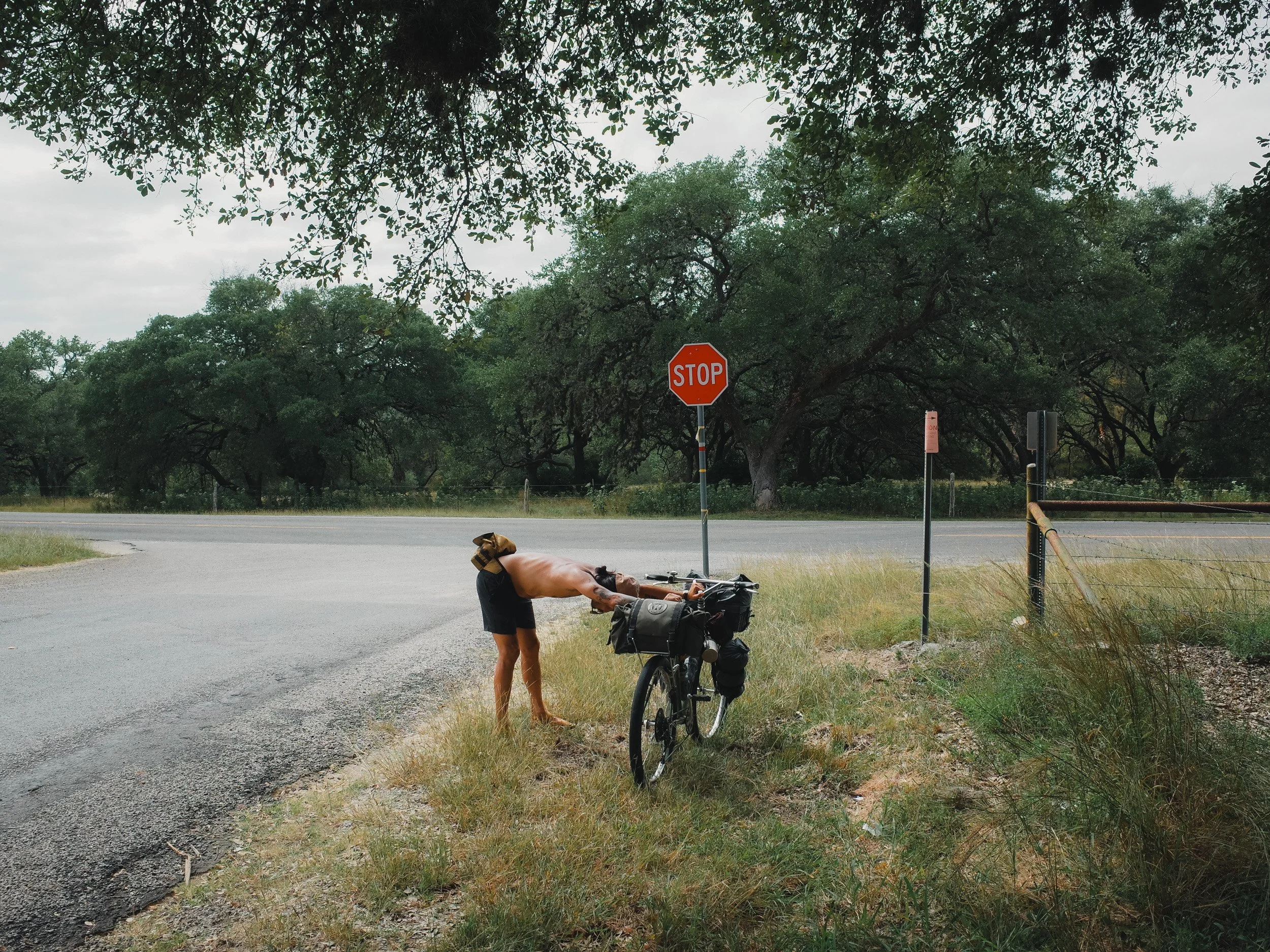 A shirtless man with backpack bending over by a bicycle near a stop sign on a rural road, surrounded by trees and grass.