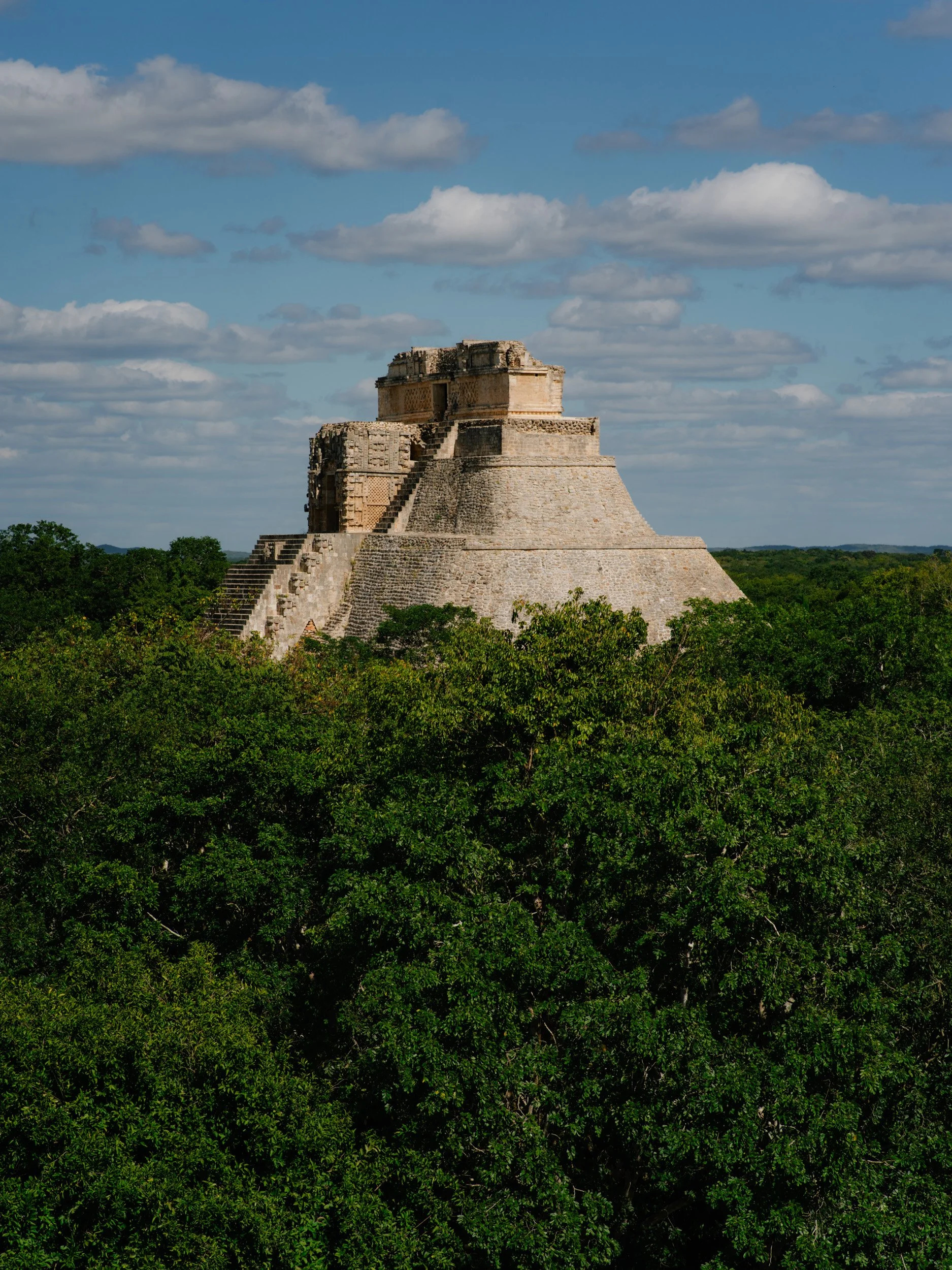 Ancient stone pyramid at Uxmal rising above dense green trees under a partly cloudy sky.