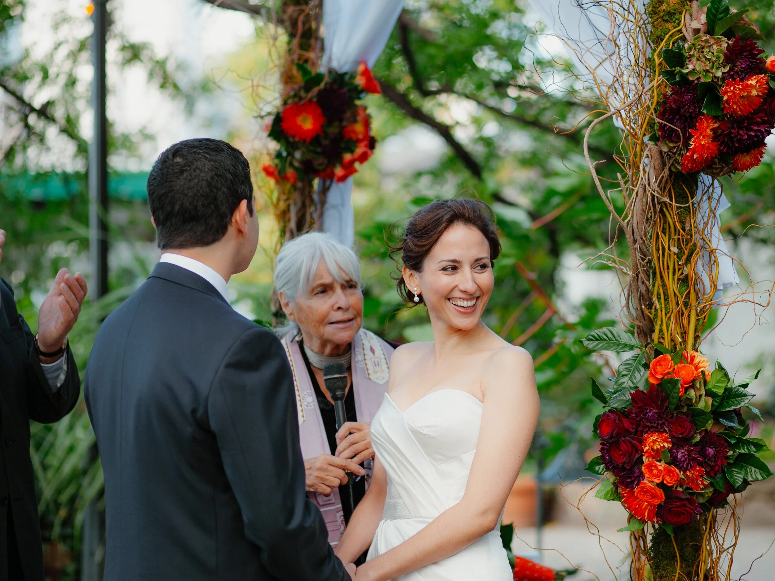 A bride and groom exchanging vows at their outdoor wedding ceremony, with a floral arch and an officiant holding a microphone in the background.