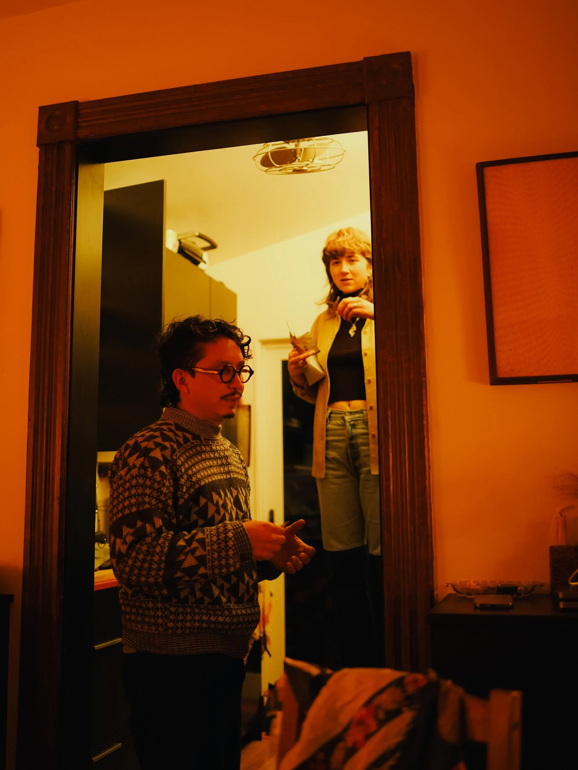 A man with glasses and a patterned sweater stands in a doorway, looking down, while a woman with short wavy hair, wearing a beige jacket and black shirt, stands on a ladder in the nearby room, holding a cup and looking out of the doorway.