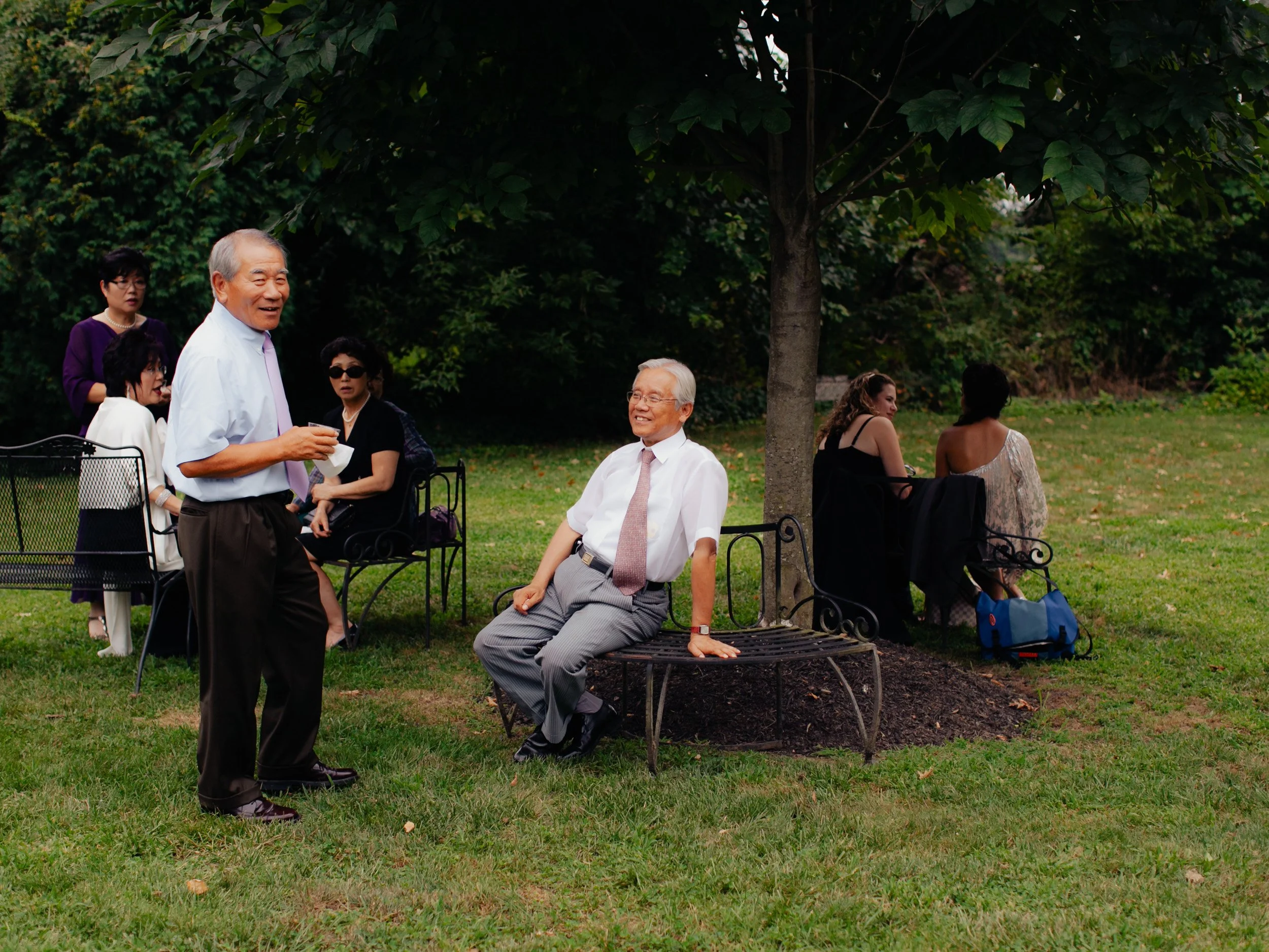 Group of seven people gathered outdoors in a park, some sitting on benches and others standing, engaging in conversation on a grassy area with trees in the background.