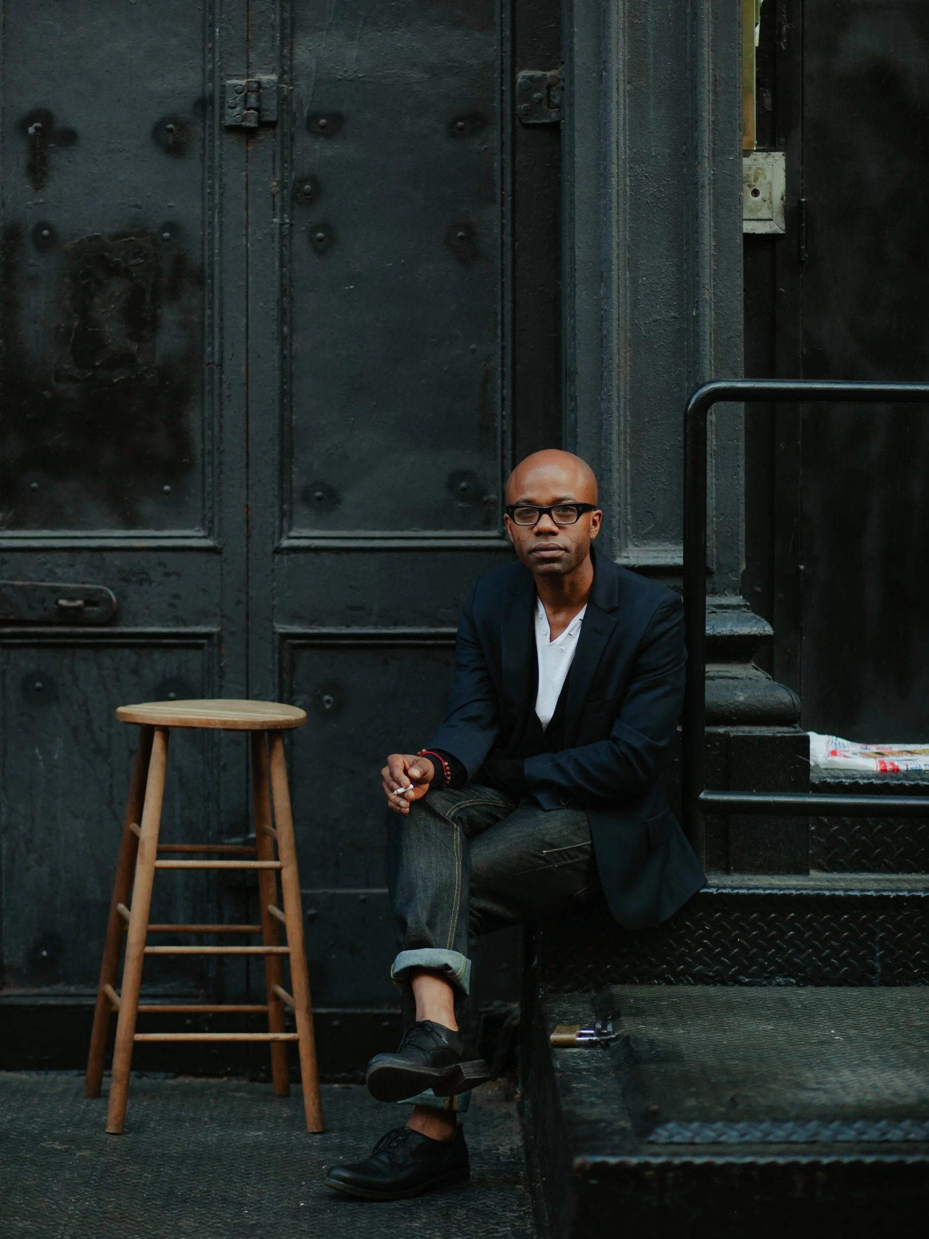 A man wearing glasses, a dark blazer, and rolled-up jeans sitting on a step outside in front of a black metal door, with a wooden stool beside him and a newspaper on the step.
