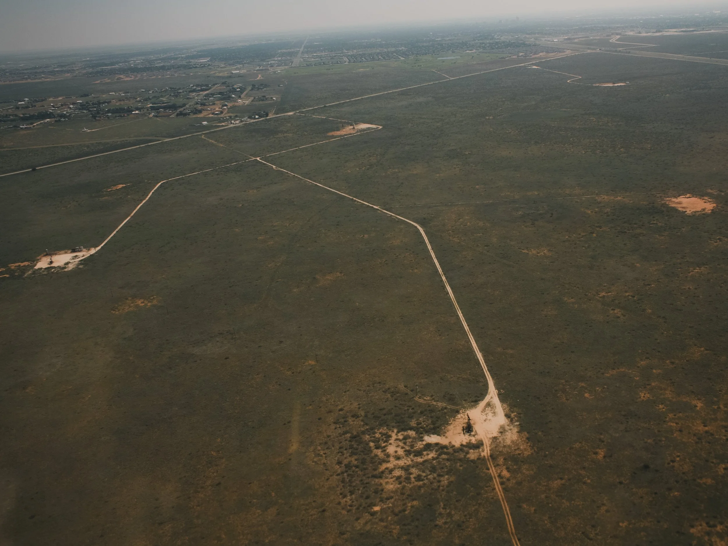 Aerial view of a large, open landscape with sparse vegetation and unpaved roads crossing through the terrain, leading to a small cleared area with an oil pump jack near the bottom right of the image and distant structures at the top.