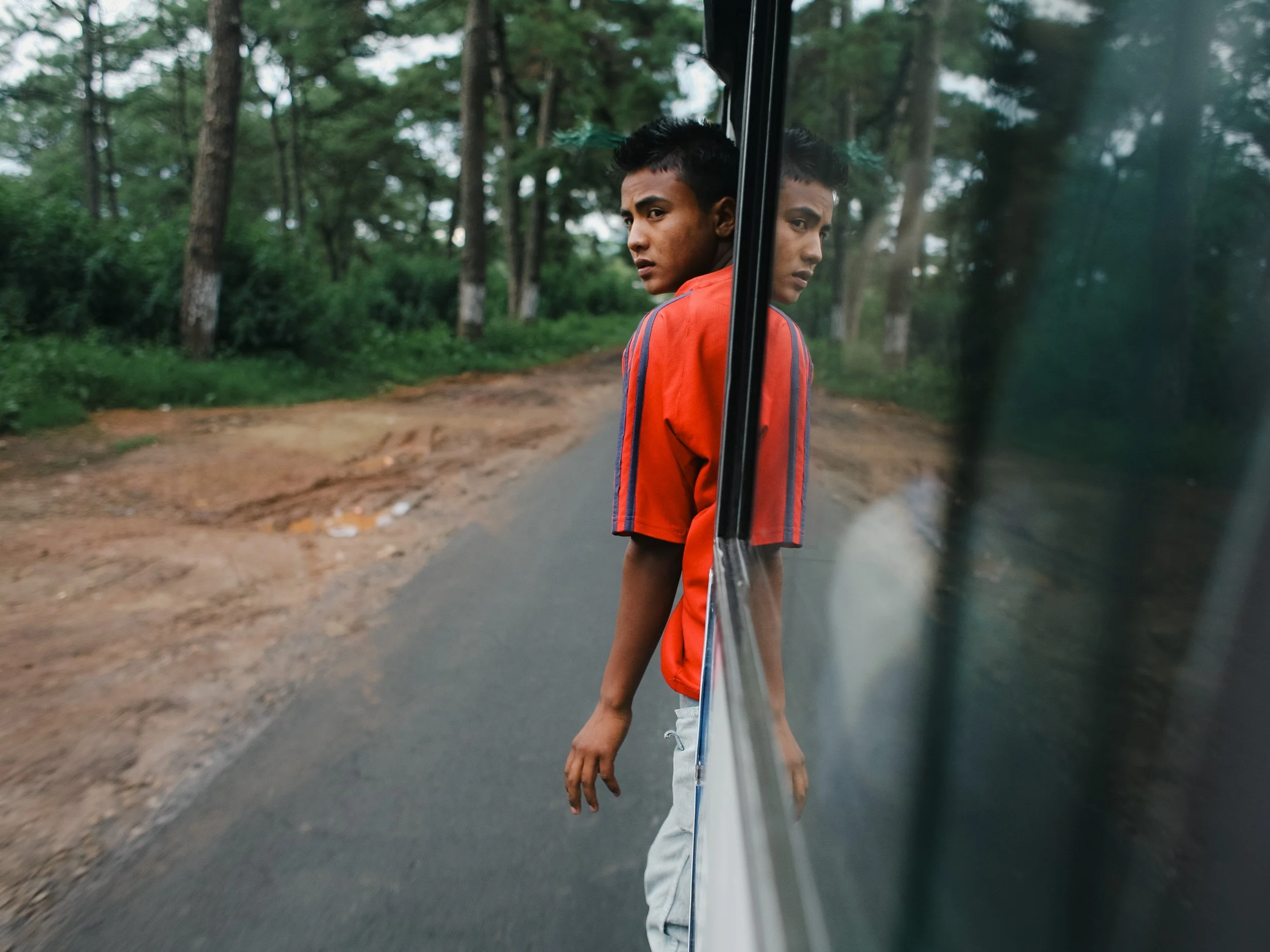 A young man in a red shirt standing outside a vehicle, with his reflection visible in the vehicle's window, on a rural road surrounded by green trees.