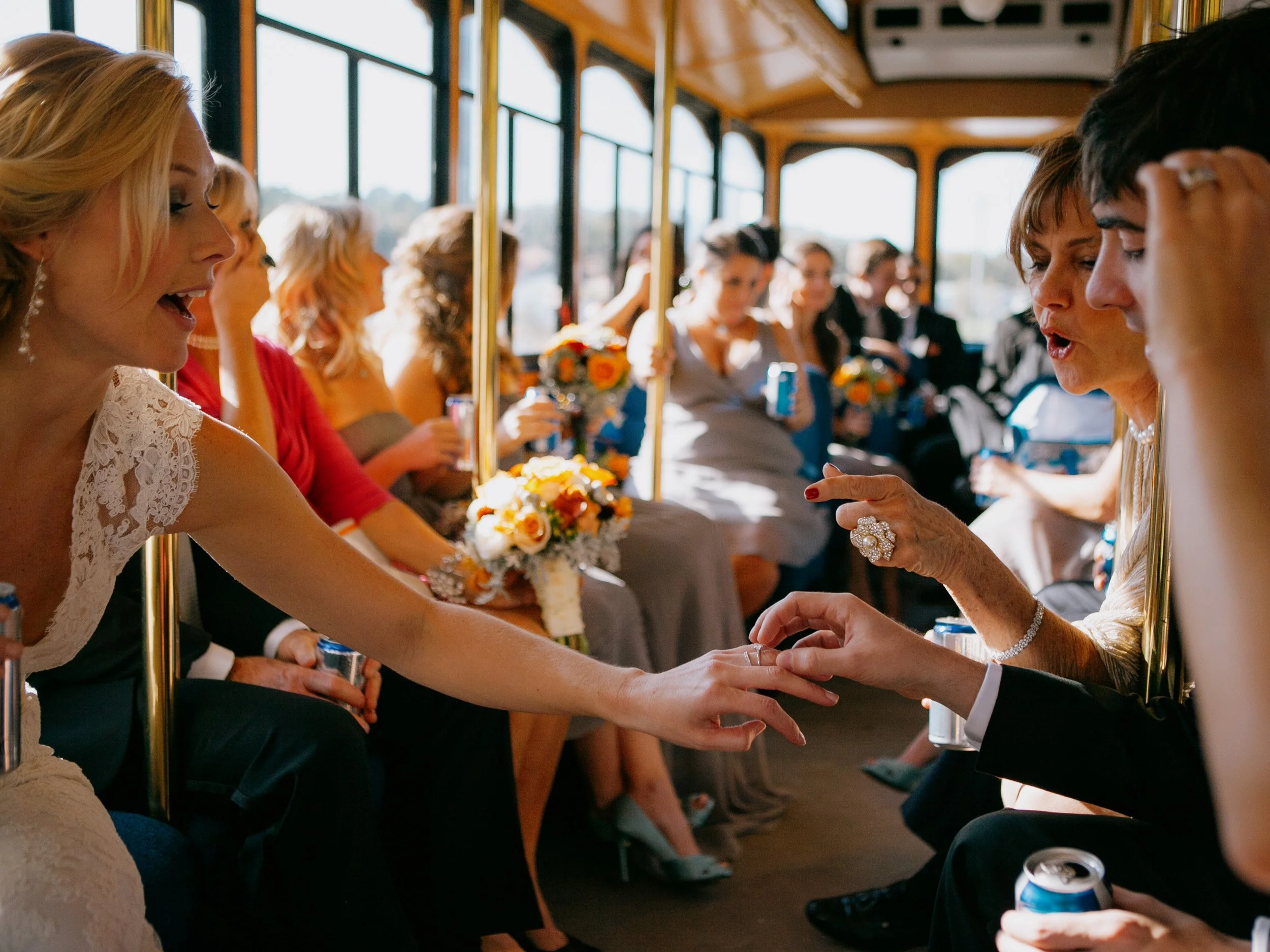 People riding on a trolley bus, with one woman touching another woman's hand, both dressed for a wedding celebration.