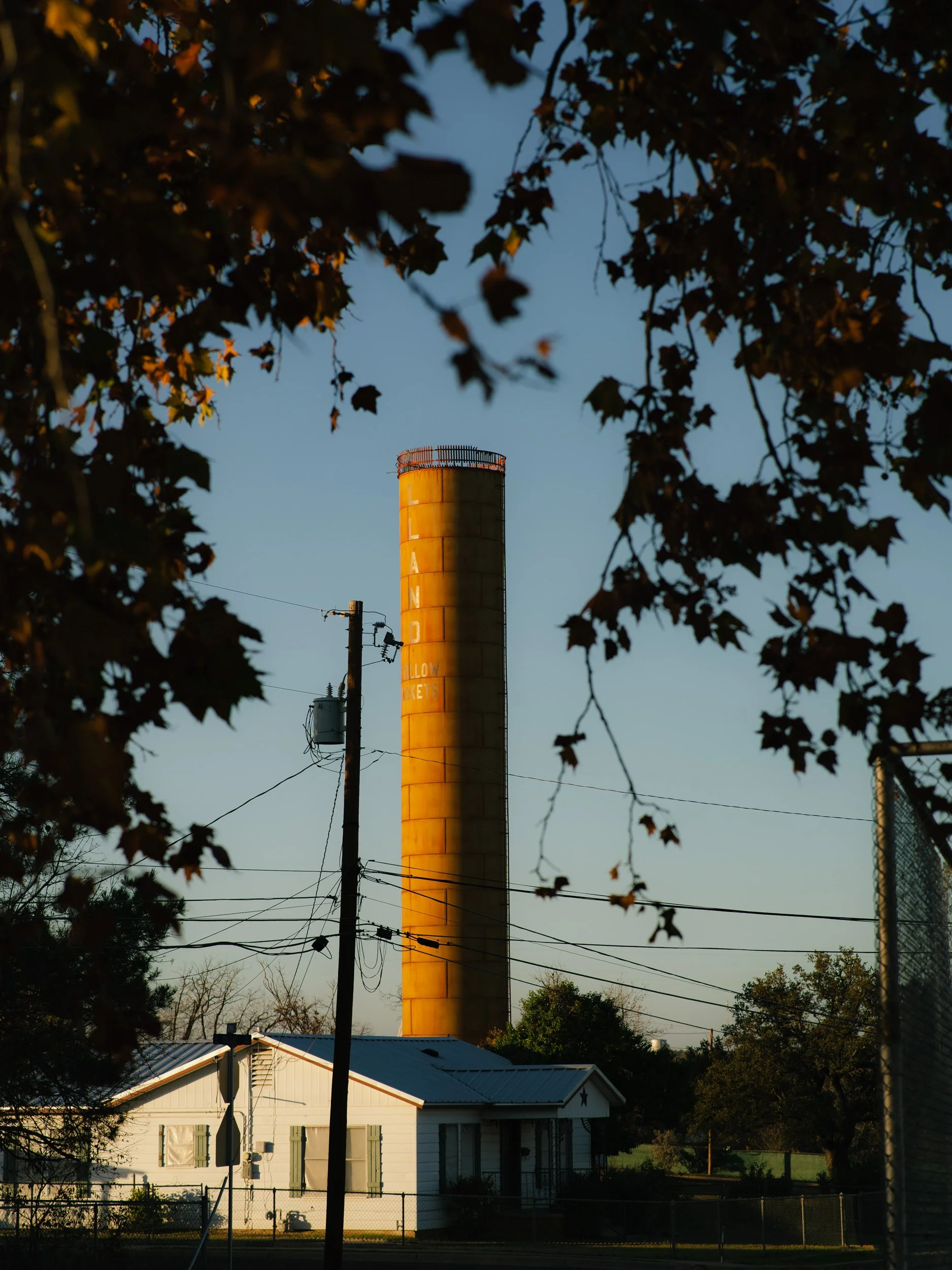 A tall industrial smokestack illuminated by sunlight, framed by tree branches and a house in the foreground.