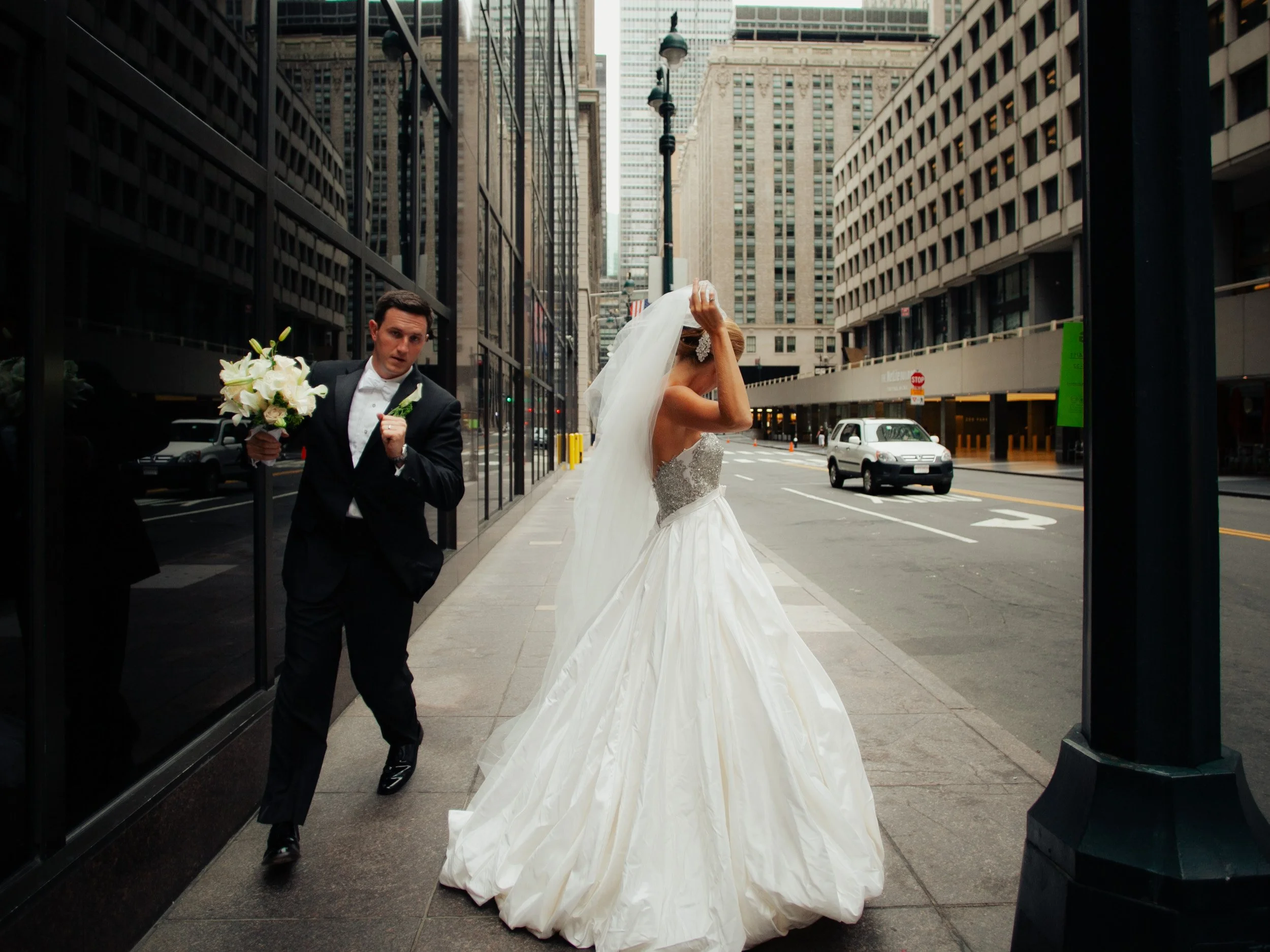 A bride in a white wedding gown and veil covers her face with her hand on a city sidewalk, while a groom in a black tuxedo holds a bouquet of white flowers and walks beside her.