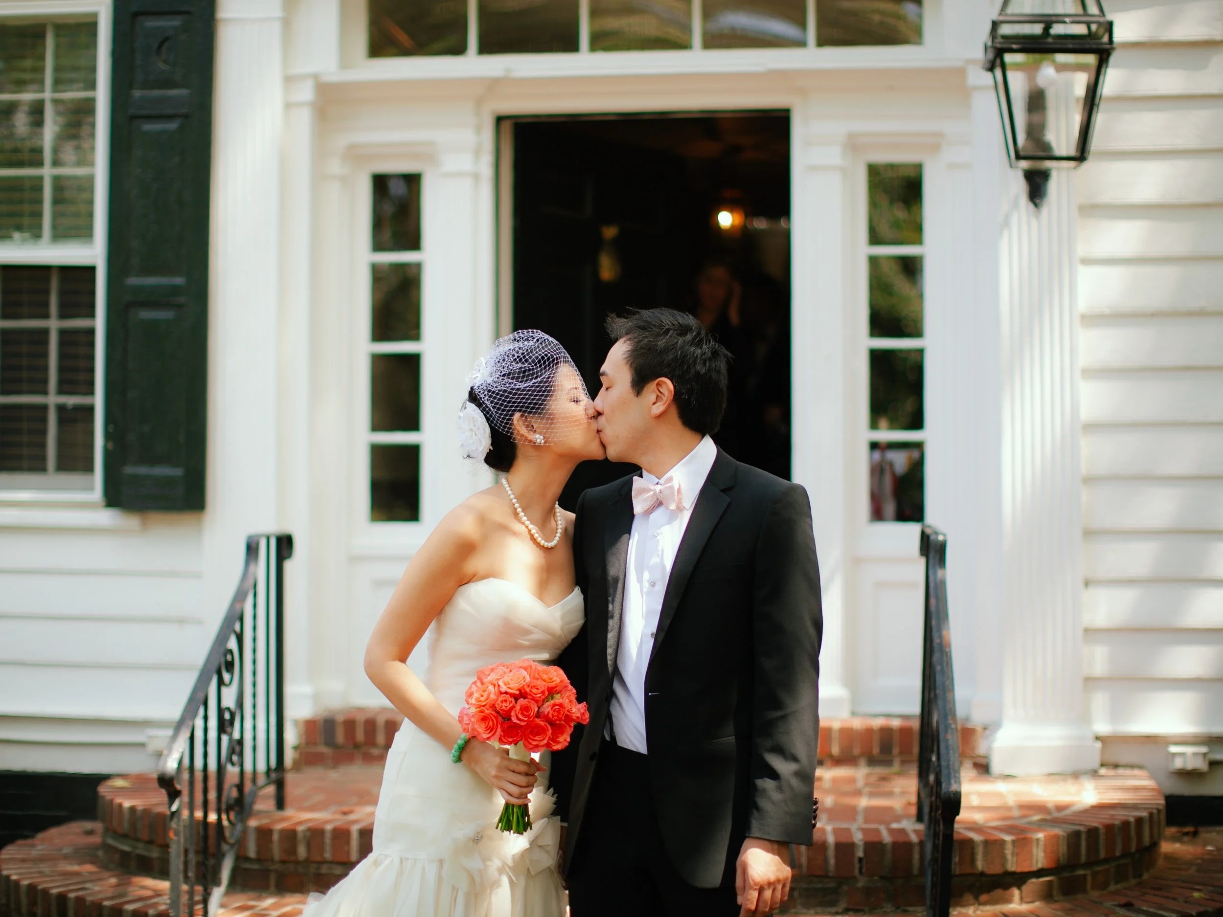 A bride and groom kissing in front of a white house, with the bride holding a bouquet of orange roses, during their wedding celebration.