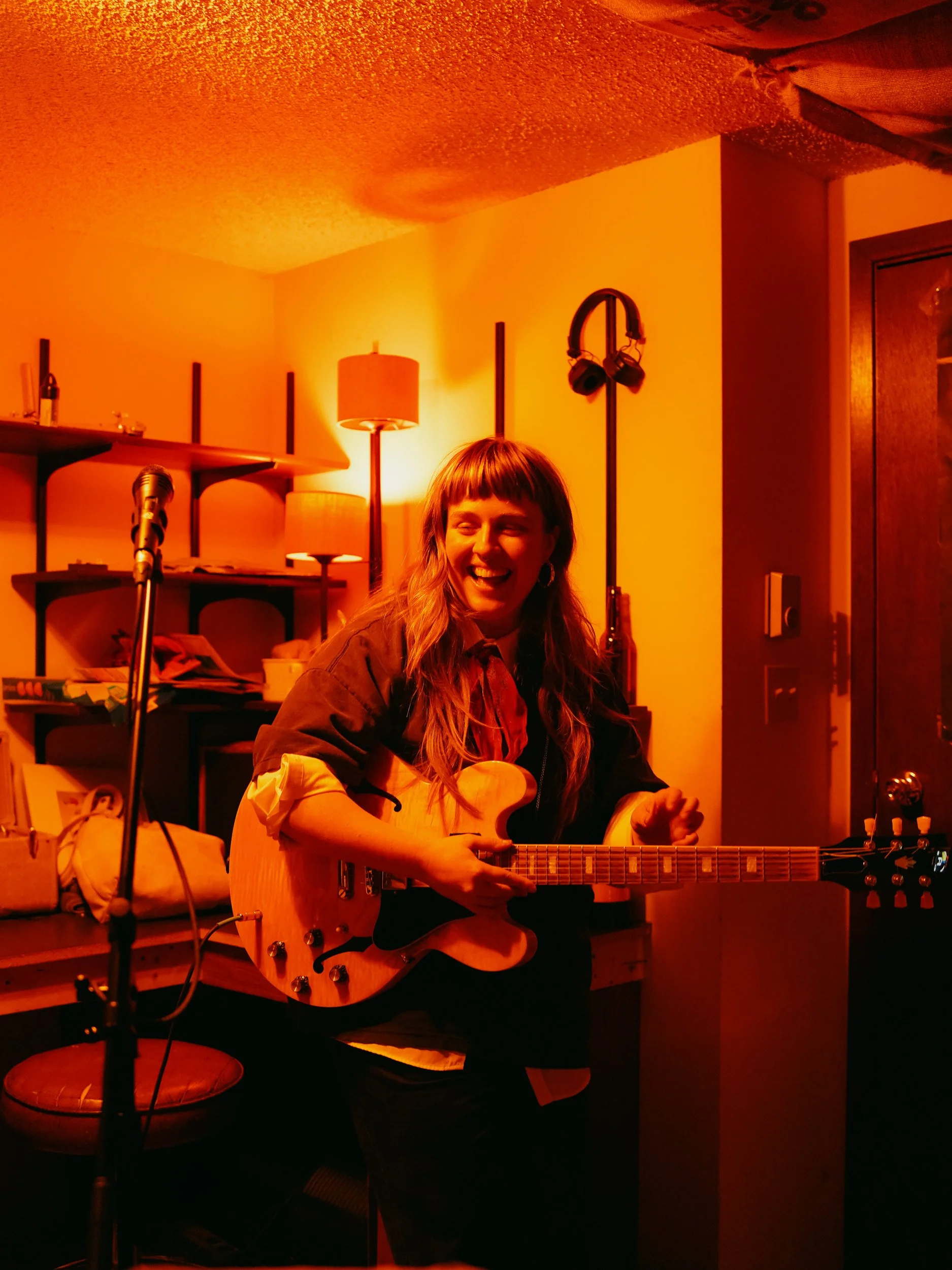 A woman laughing and playing an electric guitar in a cozy room with warm lighting, a bookshelf, and a microphone stand.