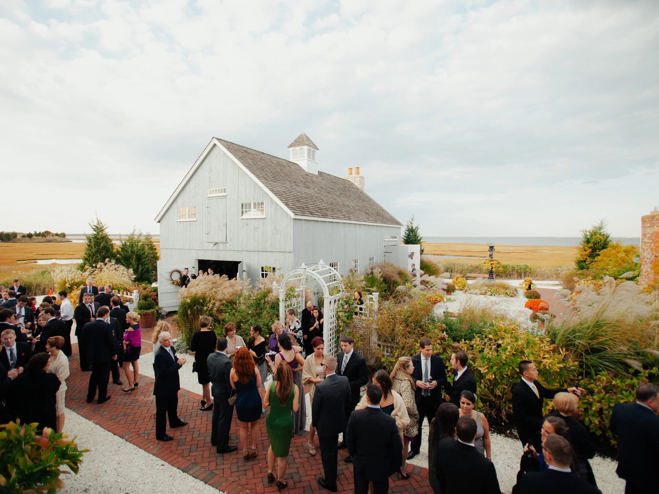 People dressed in formal attire gathering outdoors near a white barn with lush landscaping and colorful flowers on an overcast day.
