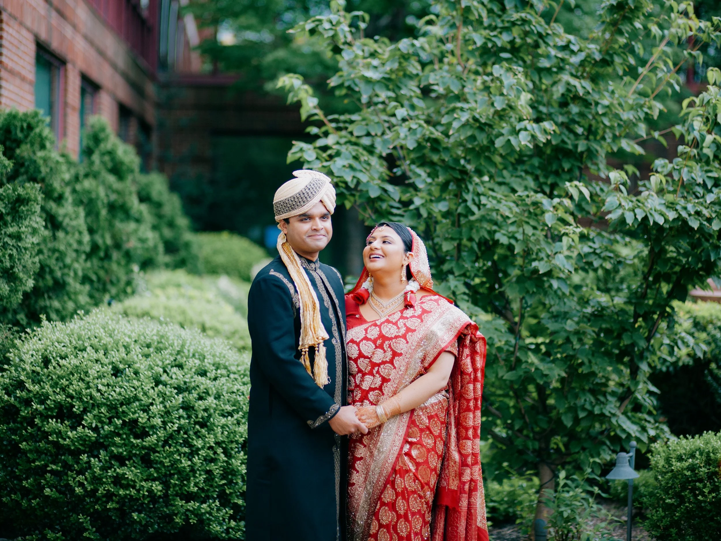 A couple dressed in traditional Indian wedding attire standing outdoors in a garden, holding hands and looking at each other, with lush green bushes and trees in the background.