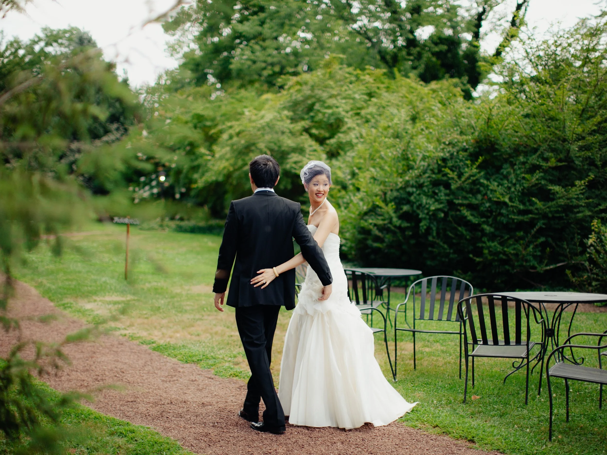 A bride and groom walking hand in hand outdoors on a garden path, surrounded by lush green trees and shrubbery, with empty black metal chairs nearby.