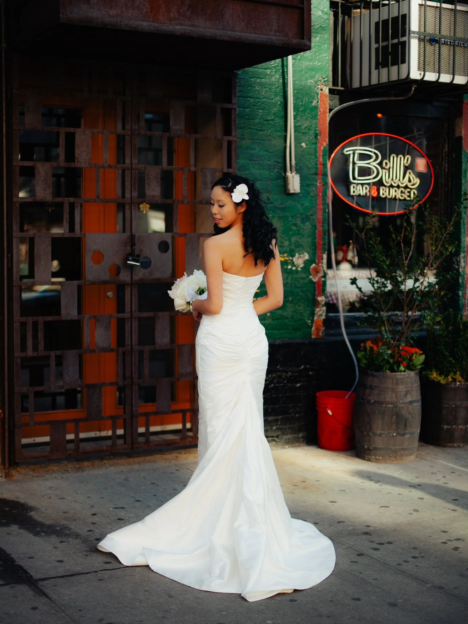 A woman dressed in a white wedding gown holding a bouquet of white flowers, standing outside near a restaurant with a neon sign that reads 'Billy's Bar & Burger' in red and white.