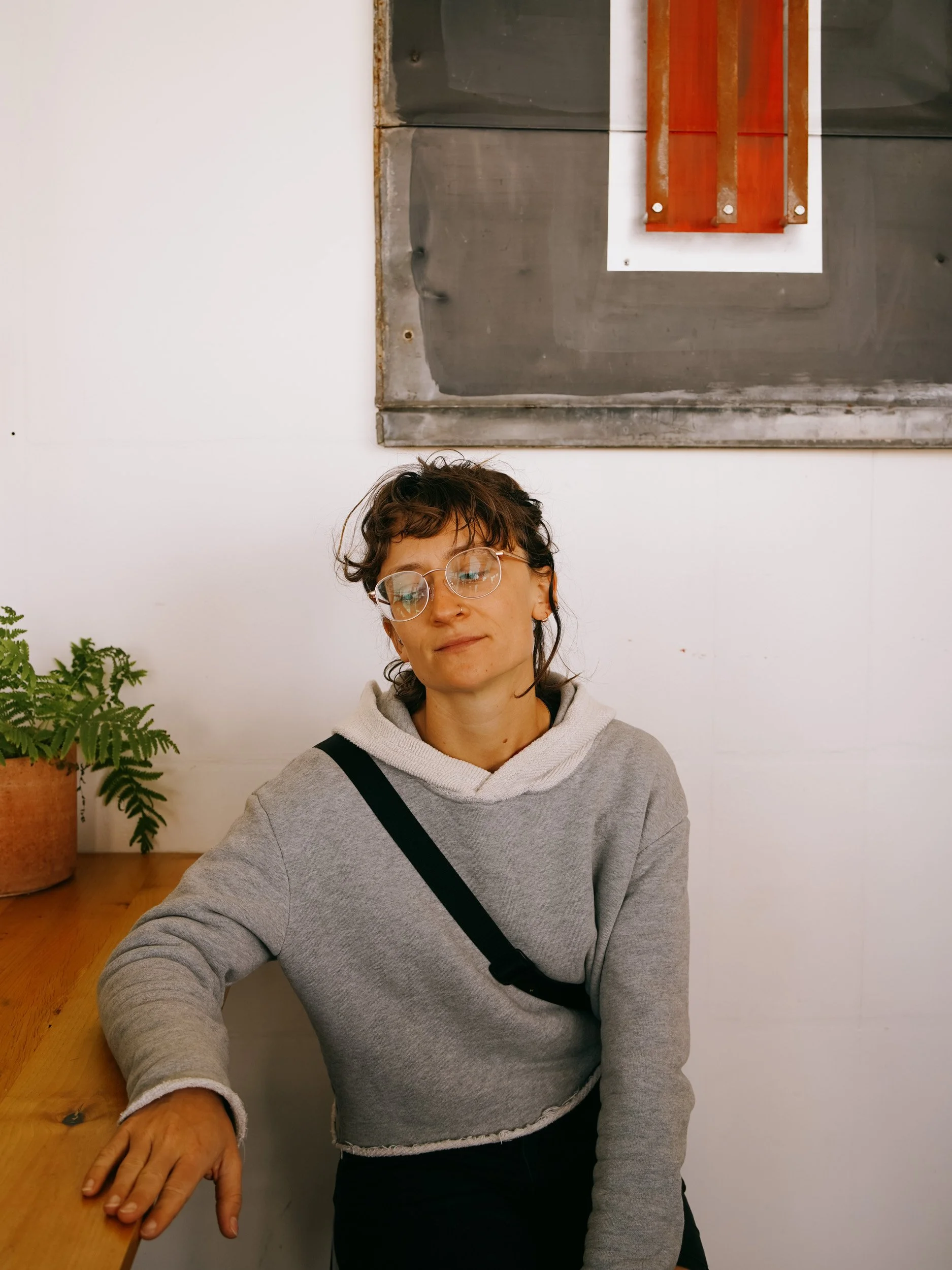 A woman with short wavy brown hair and glasses, wearing a gray hoodie with a black strap, sitting at a wooden table with her hand resting on it, in a room with white walls, a green potted plant, and a metallic and wooden decorative piece hanging on t