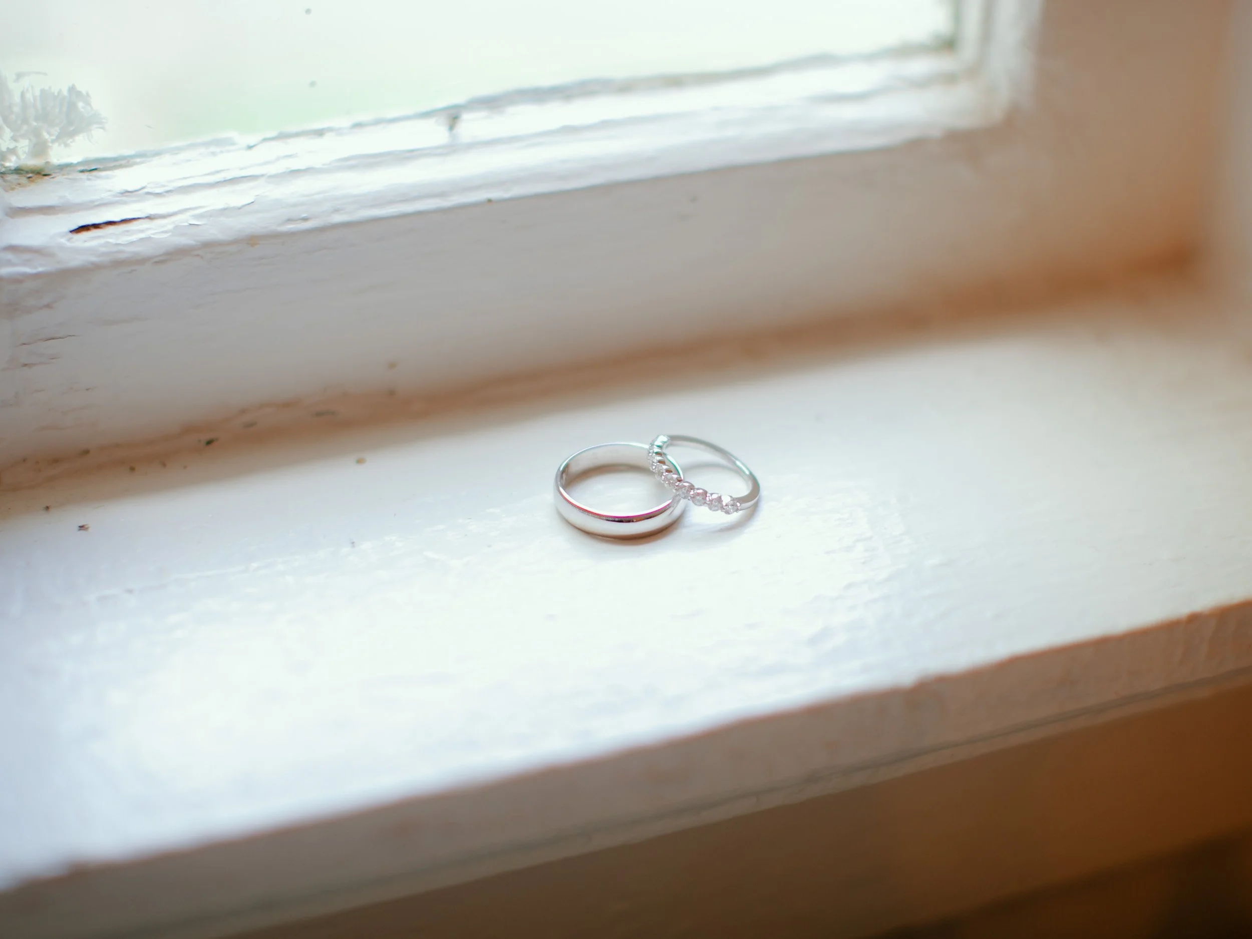 Two silver rings, one plain band and one with small gemstones, placed on a white windowsill near a window.
