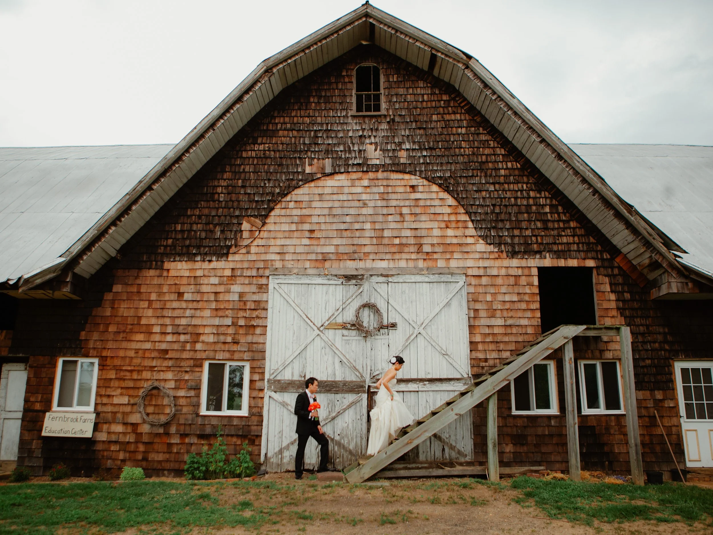 A bride and groom standing in front of a rustic barn, with the bride climbing a small wooden staircase and the groom holding a bouquet of orange flowers.