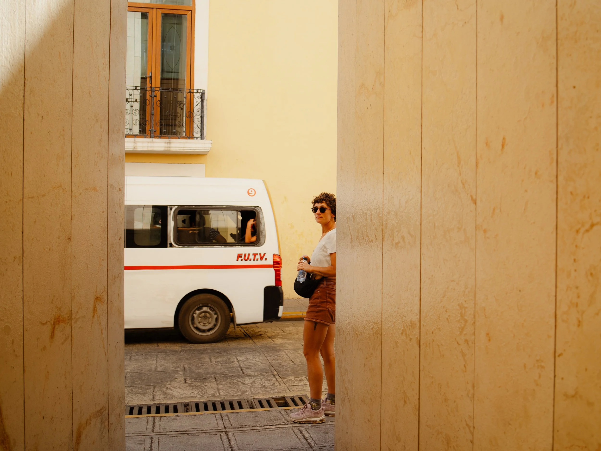 A woman with sunglasses and casual clothing standing in a narrow alleyway between large yellow walls, with a white van with red stripe and letters parked behind her.
