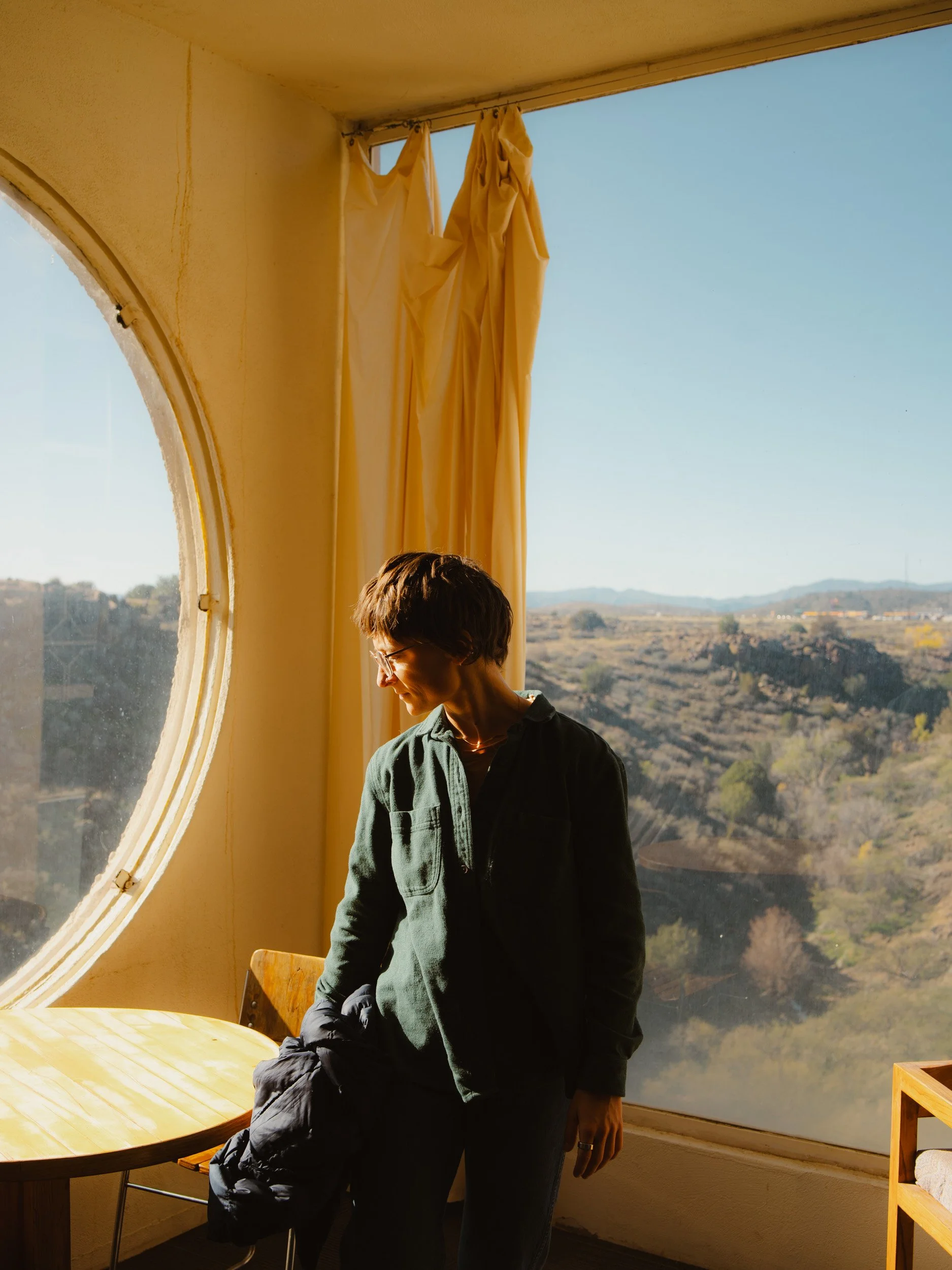 A person with short hair and glasses standing by a window with a mountain view, warm sunlight coming through, in a room with cream-colored curtains and a wooden table.