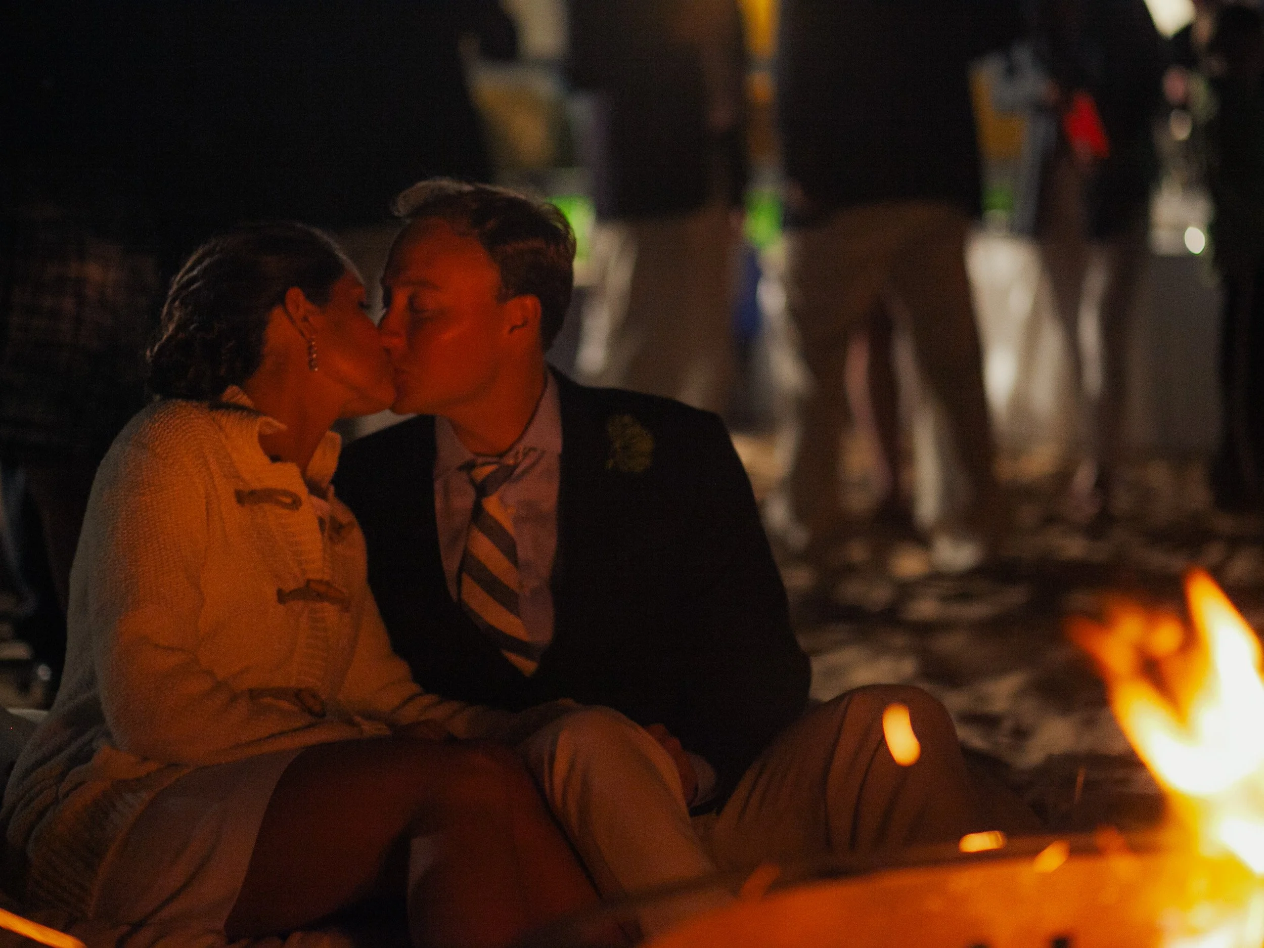 A couple kissing by a campfire at night with people in the background.