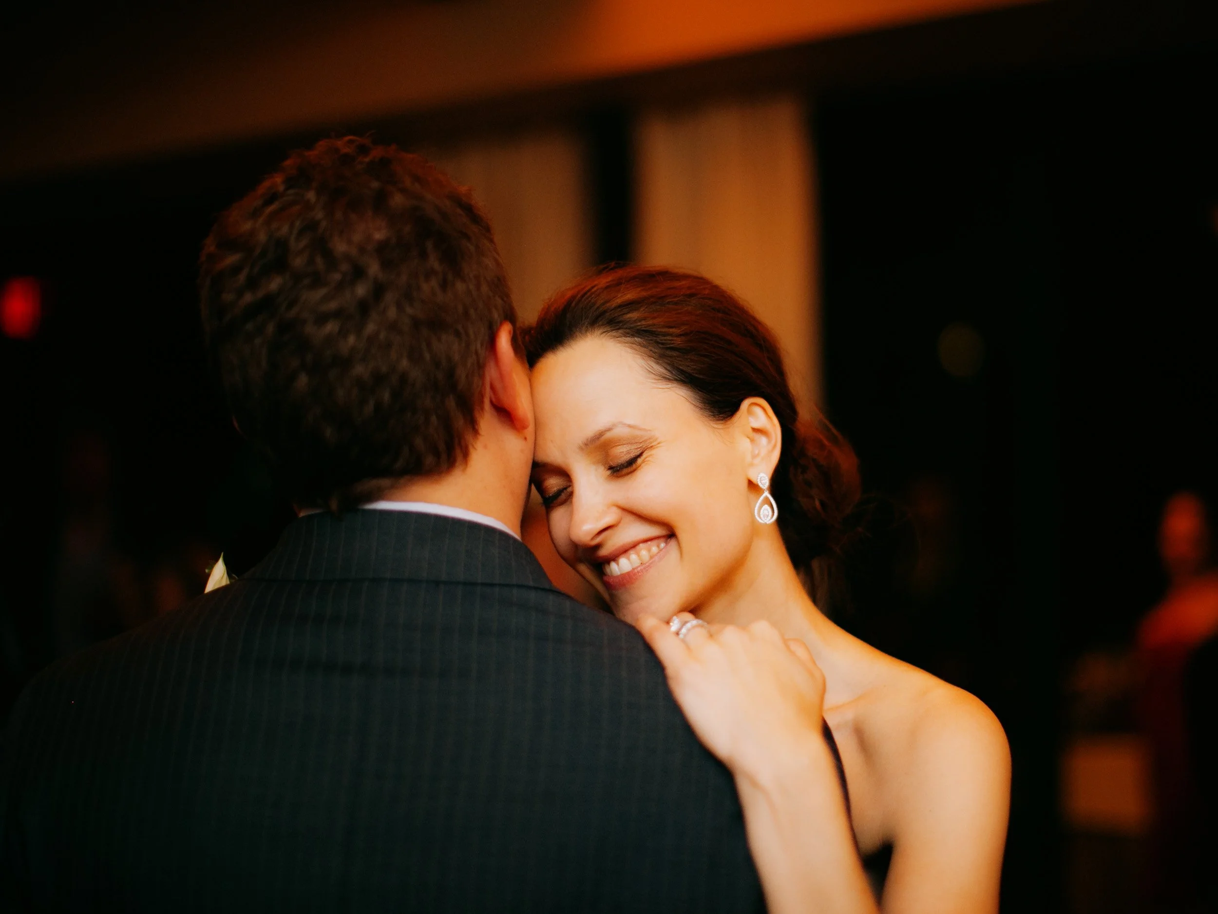 A woman smiling with her eyes closed, dance partner's back visible, in a dimly lit room at a dance or wedding event.