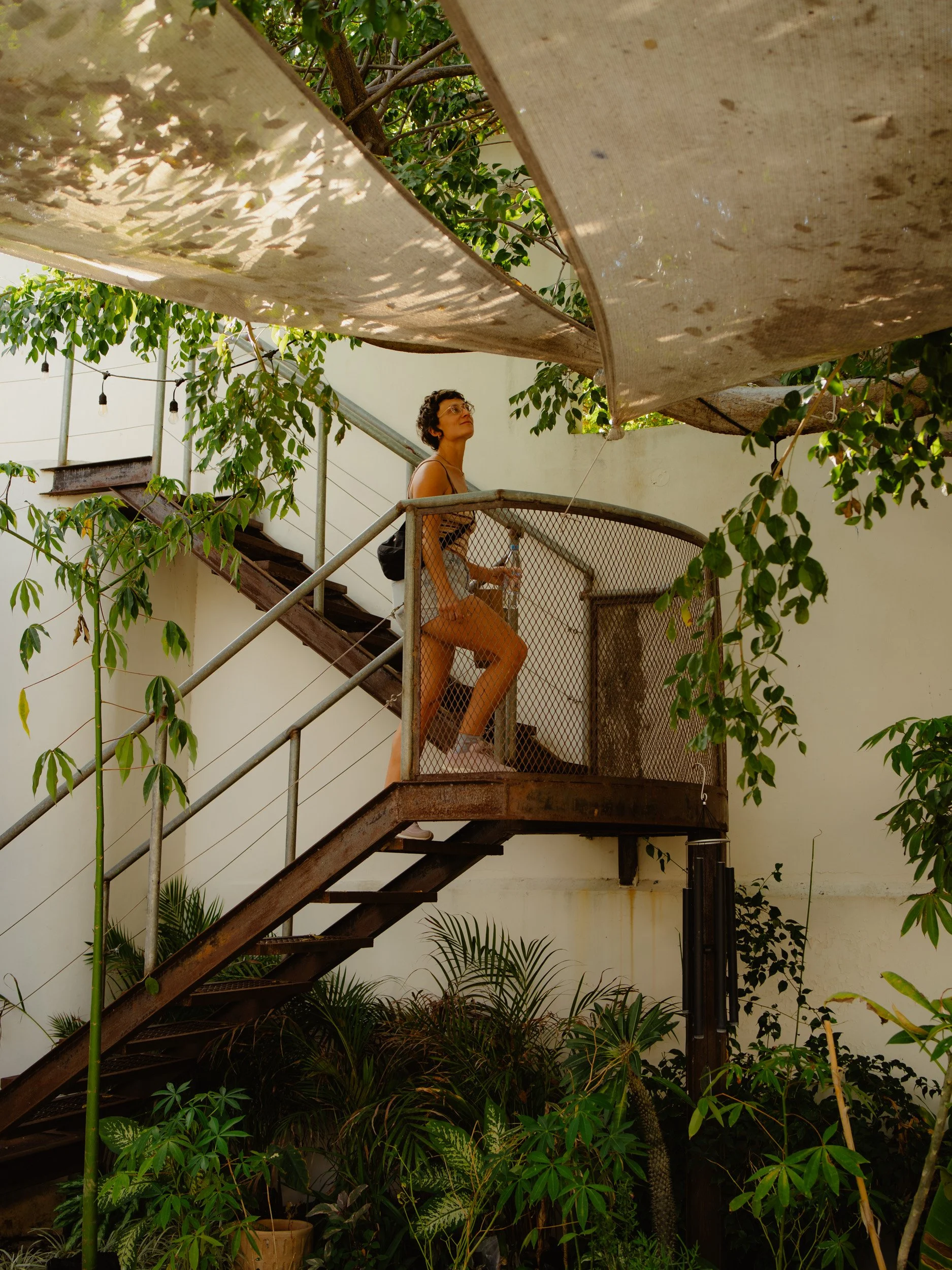 A woman standing on a metal staircase and balcony outdoors, surrounded by green plants and trees, with a white wall in the background. She is wearing glasses, a tank top, and shorts, and holding a water bottle.