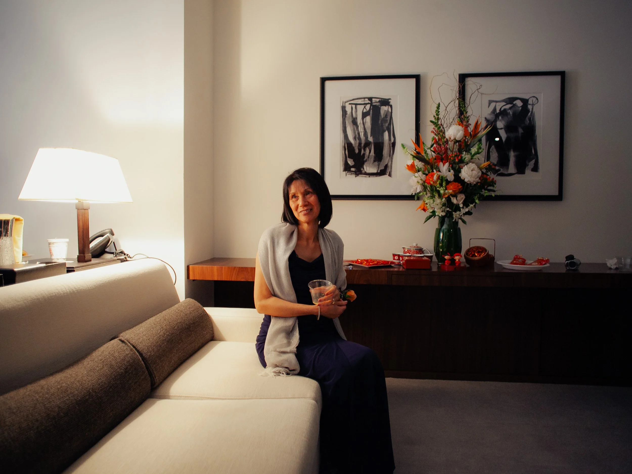 Woman sitting on a beige sofa in a hotel room, holding a drink and smiling. There is a lamp and a phone on the side table behind her, and framed black-and-white artwork and a large floral arrangement on the back wall.