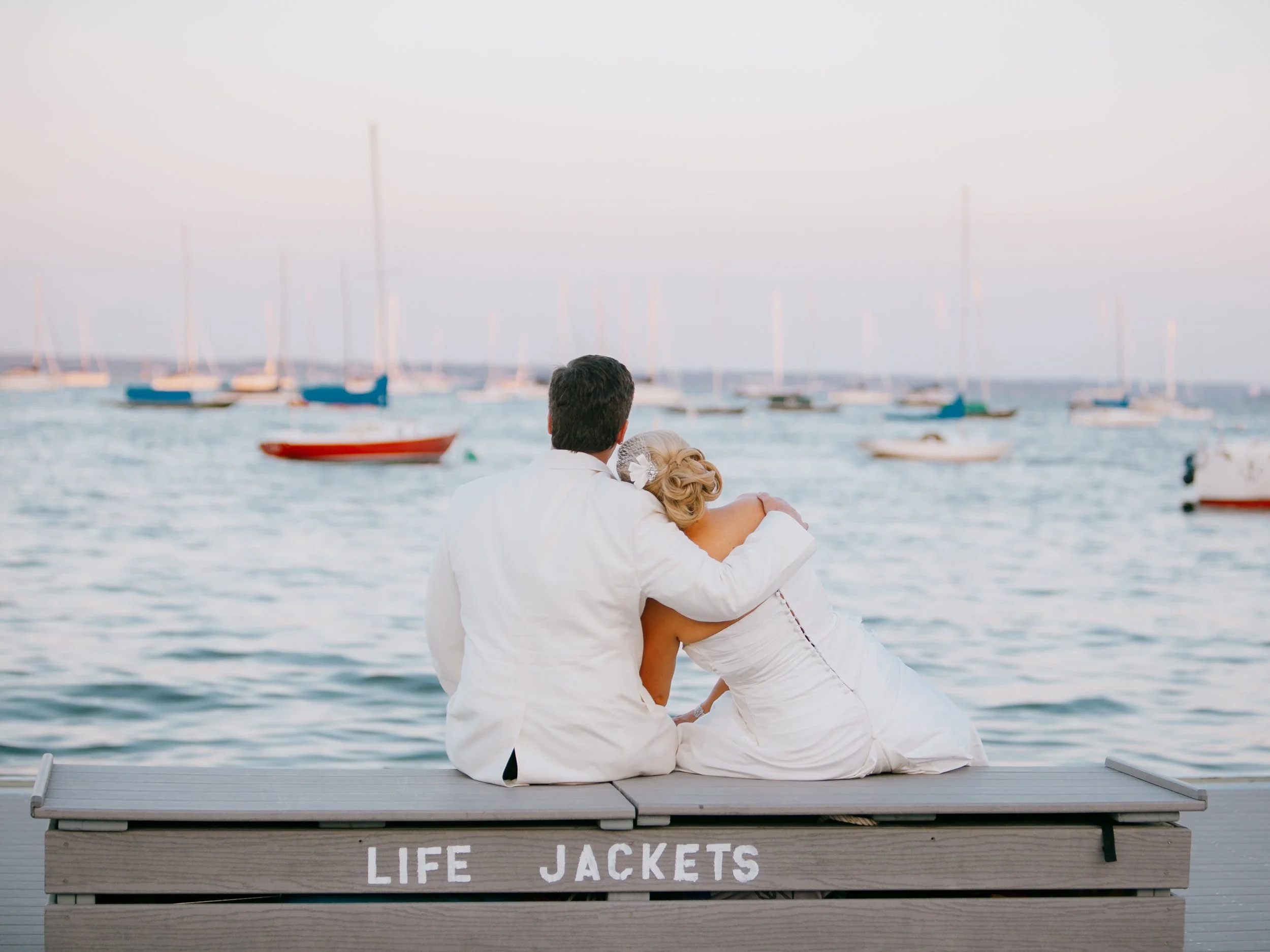 A couple in wedding attire sitting on a bench labeled 'LIFE JACKETS' by the water, looking at sailboats on the ocean during sunset.