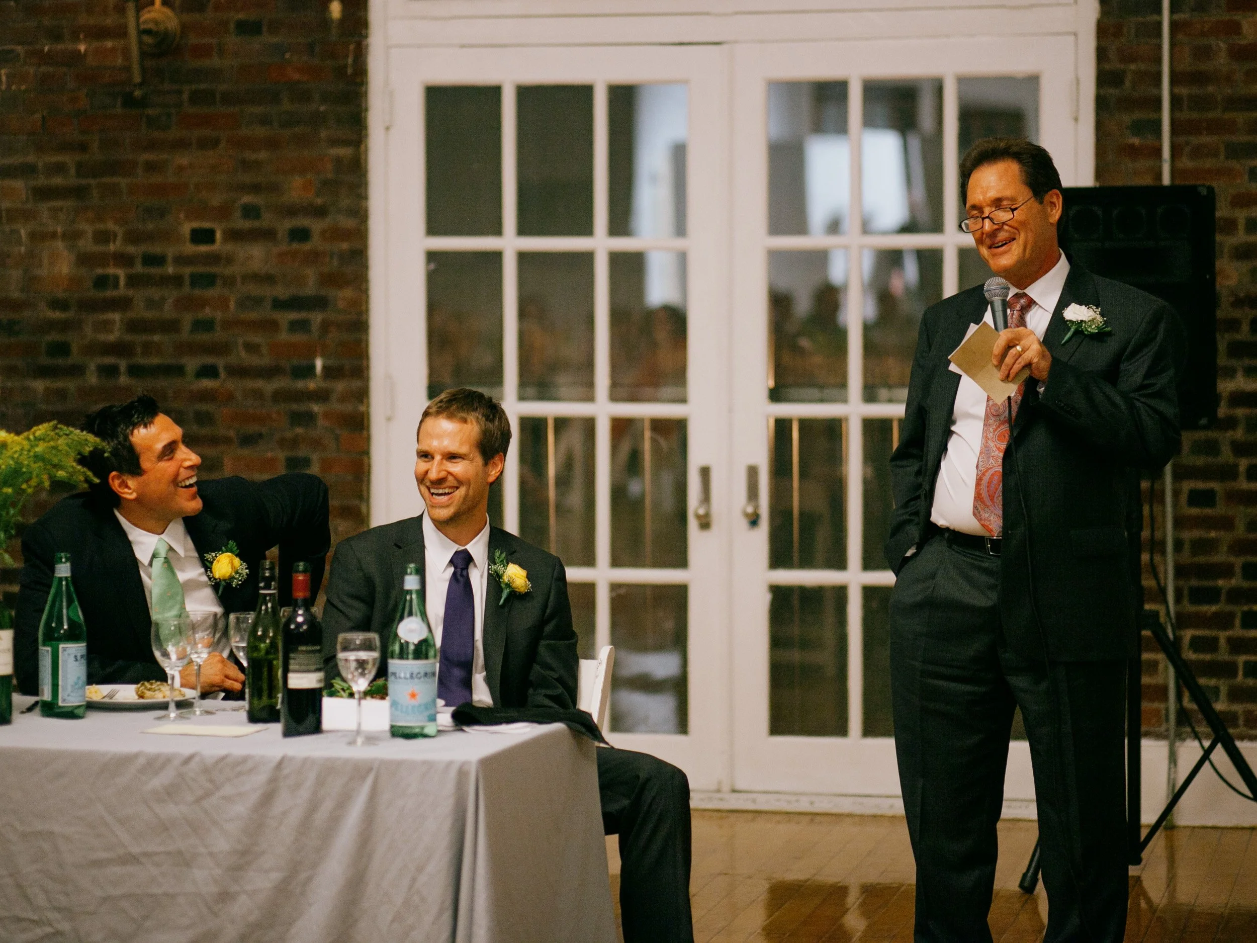 A wedding reception scene with two men sitting at a table, smiling and laughing, and an older man standing nearby giving a speech, holding a microphone. The room has brick walls and large glass doors in the background.