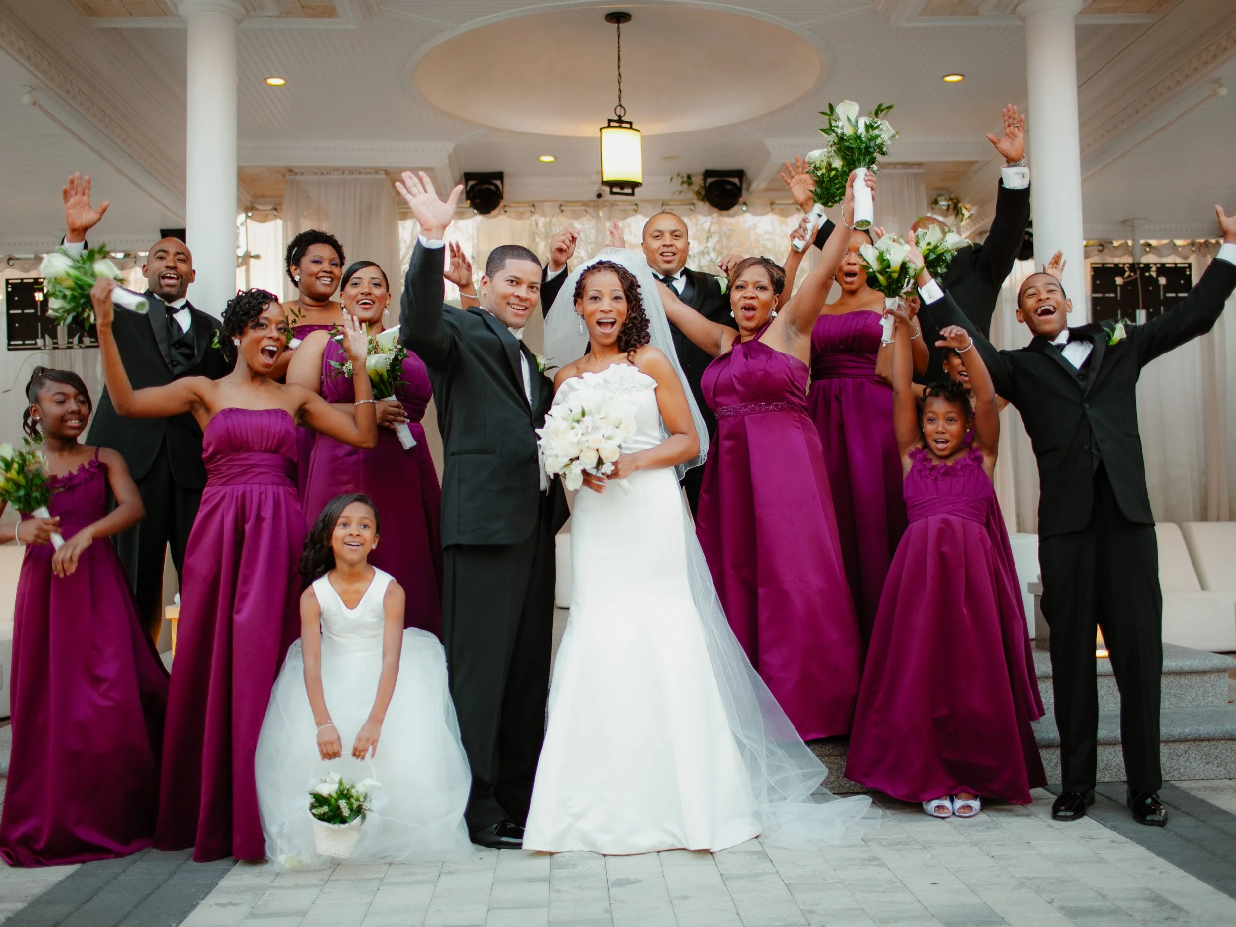 A wedding celebration with the bride and groom in the center, surrounded by family and friends, some holding bouquets, all smiling and raising their hands in joy.