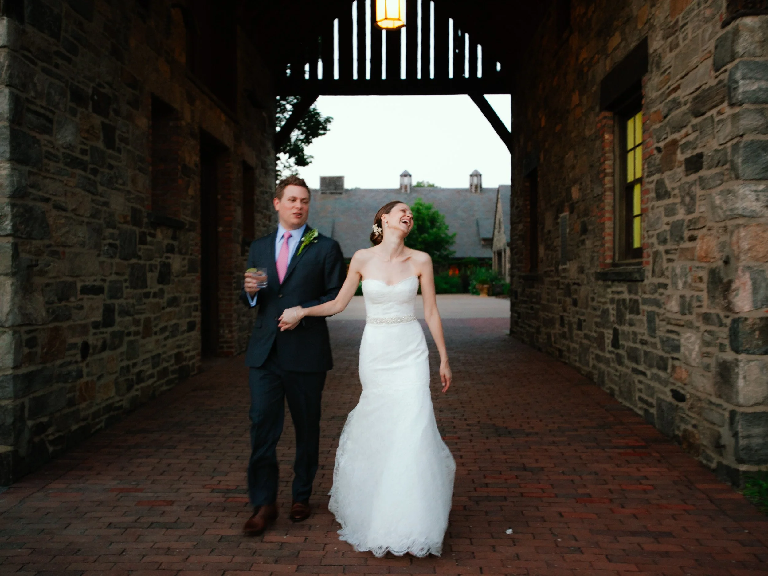 A bride and groom walking together under a stone archway at a wedding venue, smiling and holding hands.