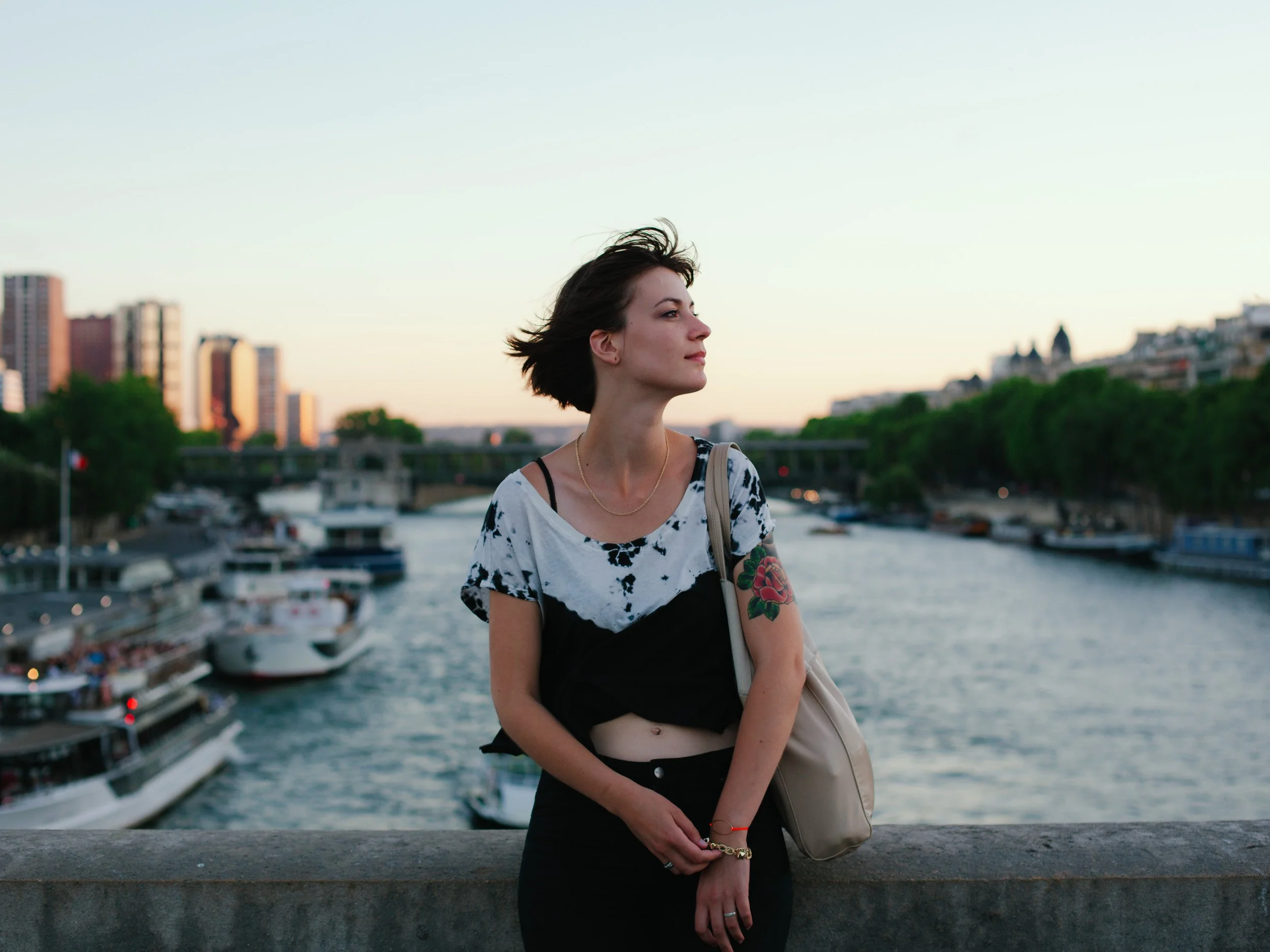 A young woman with short dark hair and tattoos on her left arm stands by a river at sunset, looking to the side with a cityscape and boats in the background.