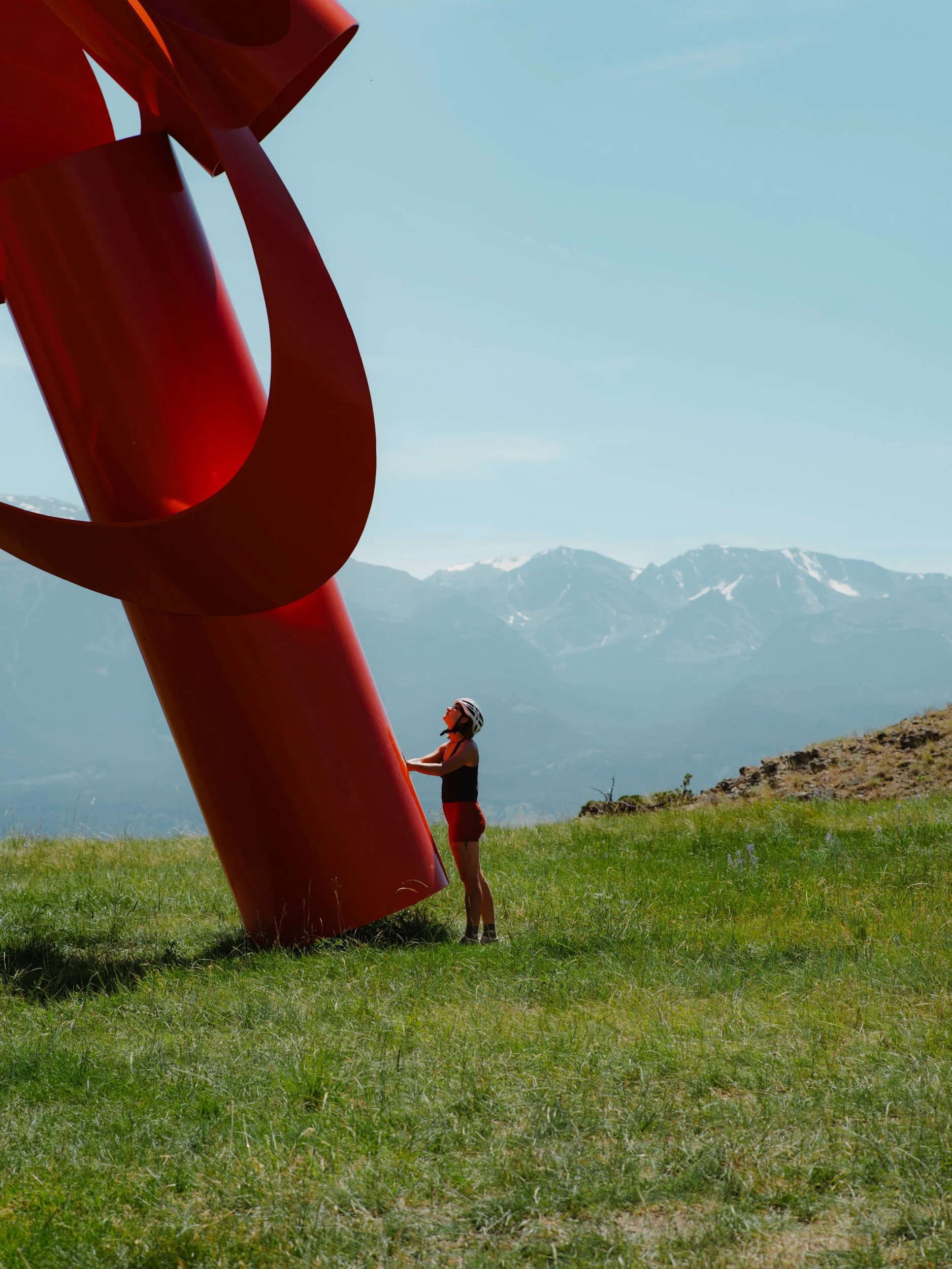 A person wearing a helmet and athletic clothing standing on grass and holding a large red sculpture with mountains in the background.