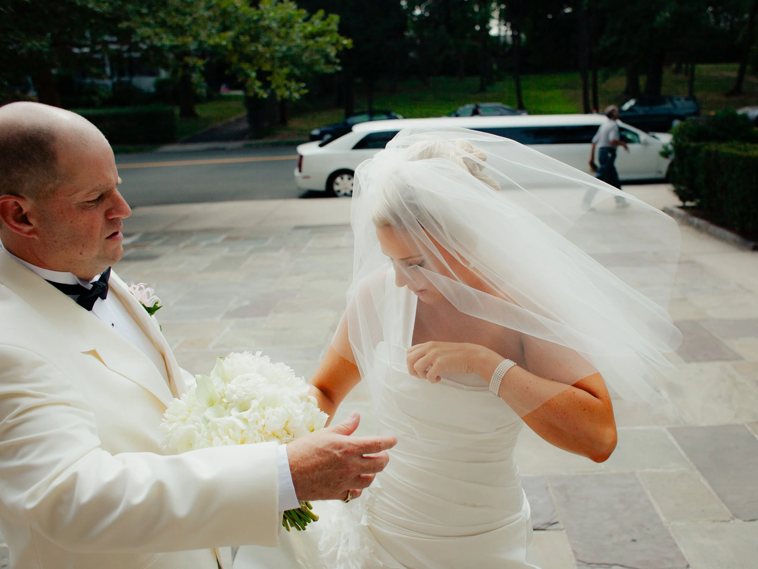 A bride with a long white veil and strapless wedding dress is looking at her hand while a man in a white tuxedo holds a bouquet of white flowers beside her outside in a parking lot.