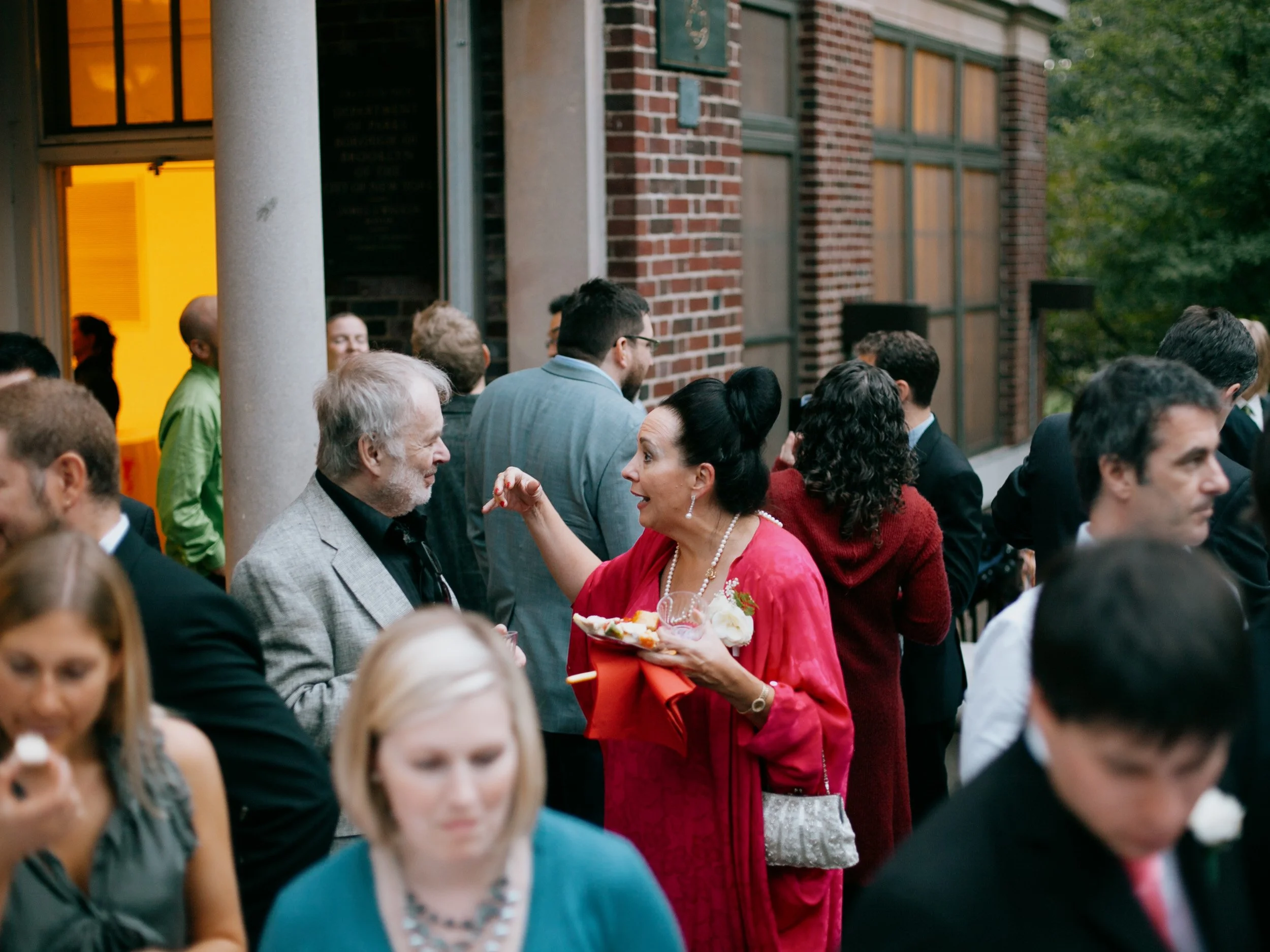 People socializing at an outdoor event, with a woman in a bright red dress talking to an older man in a gray blazer.