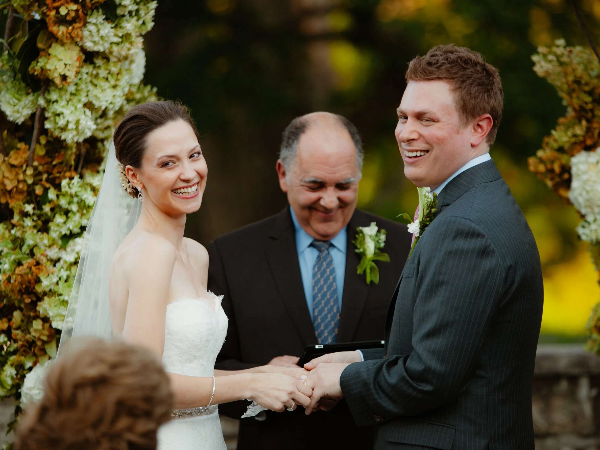 A bride and groom exchange vows at an outdoor wedding ceremony with a floral arch behind them, accompanied by an officiant.