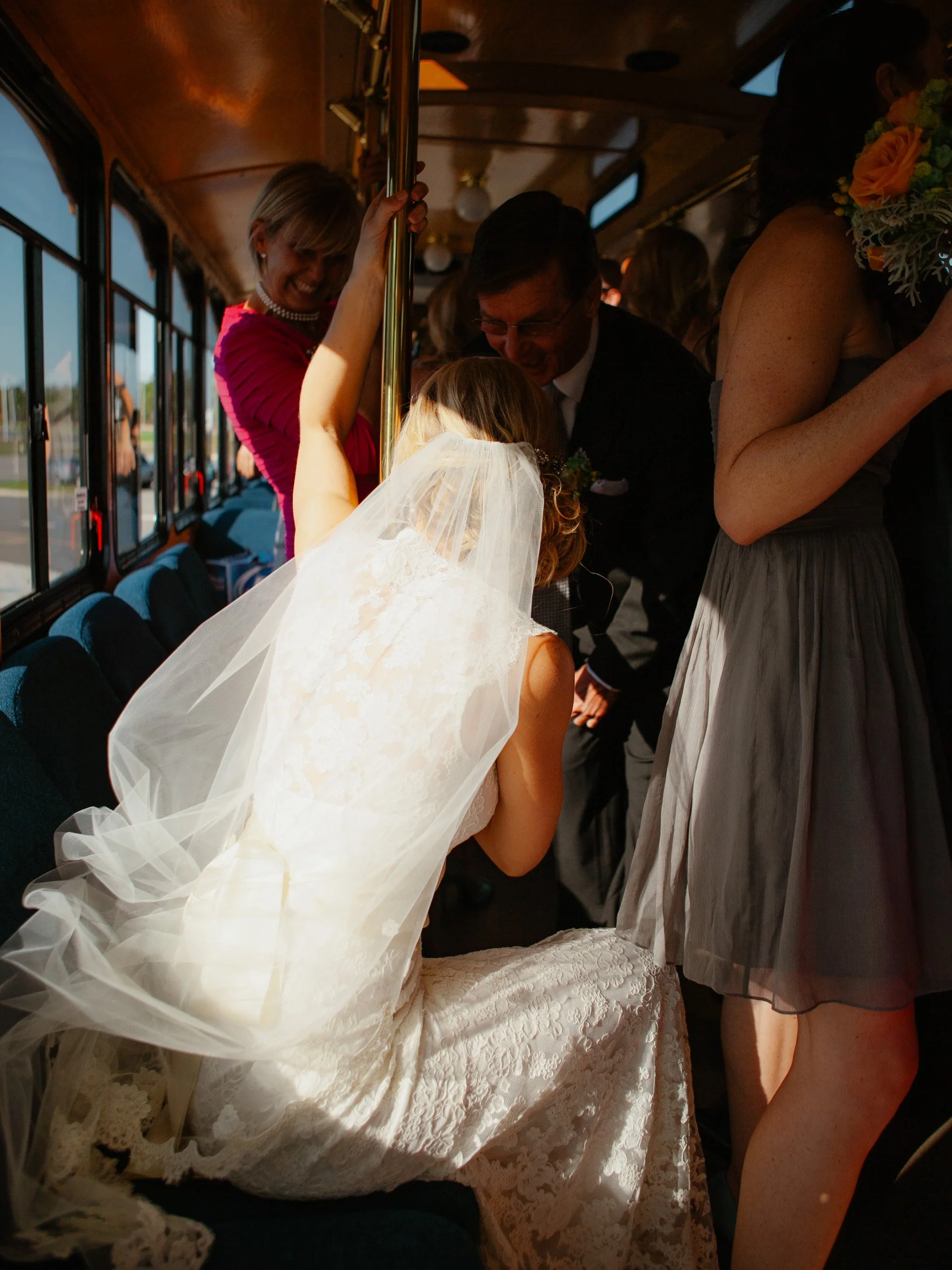 Bride sitting inside a trolley, wearing a lace wedding dress and veil, surrounded by happy guests.