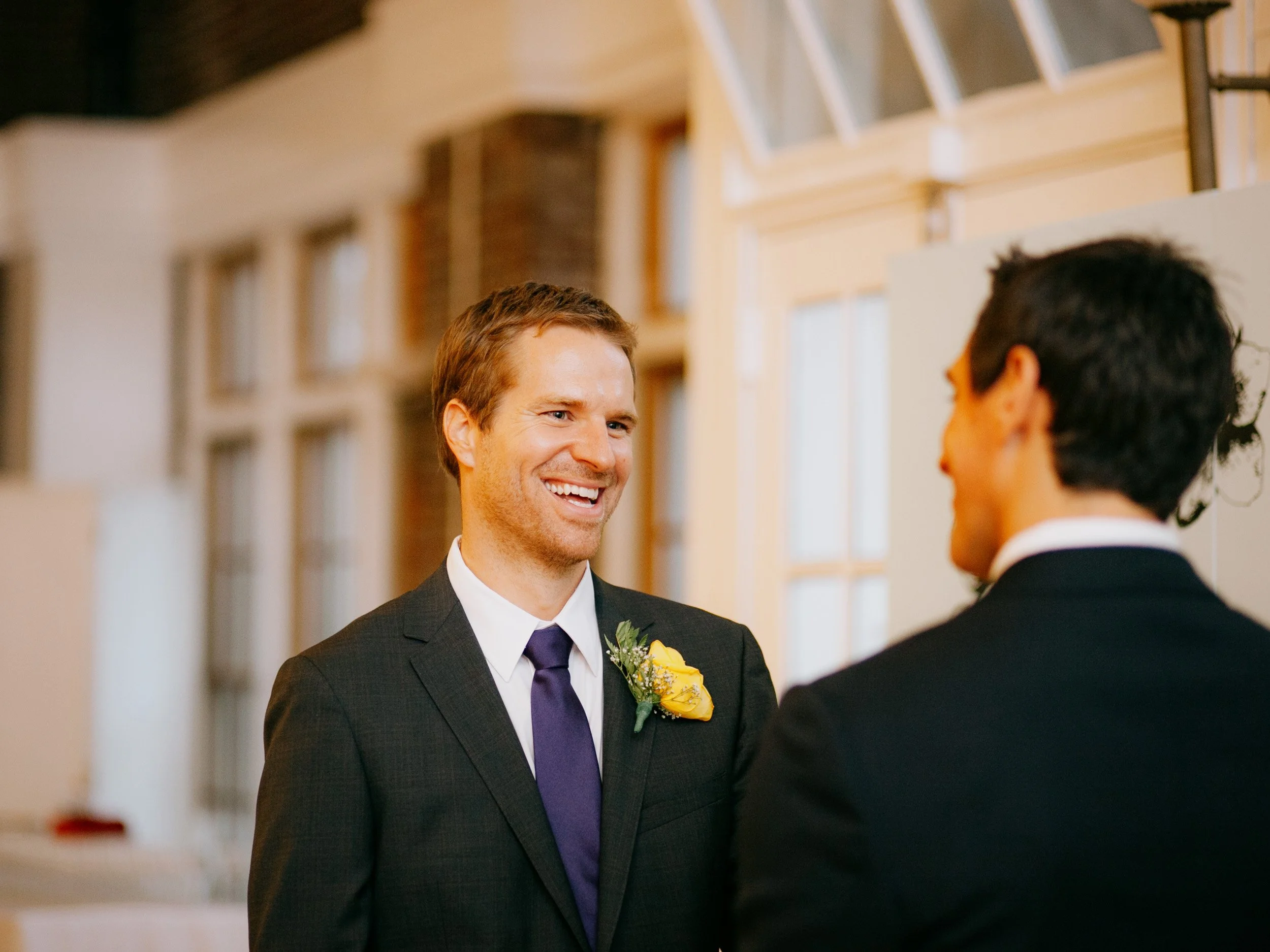 Two men in suits smiling and talking to each other indoors, with a bright, modern background featuring windows and brick walls.
