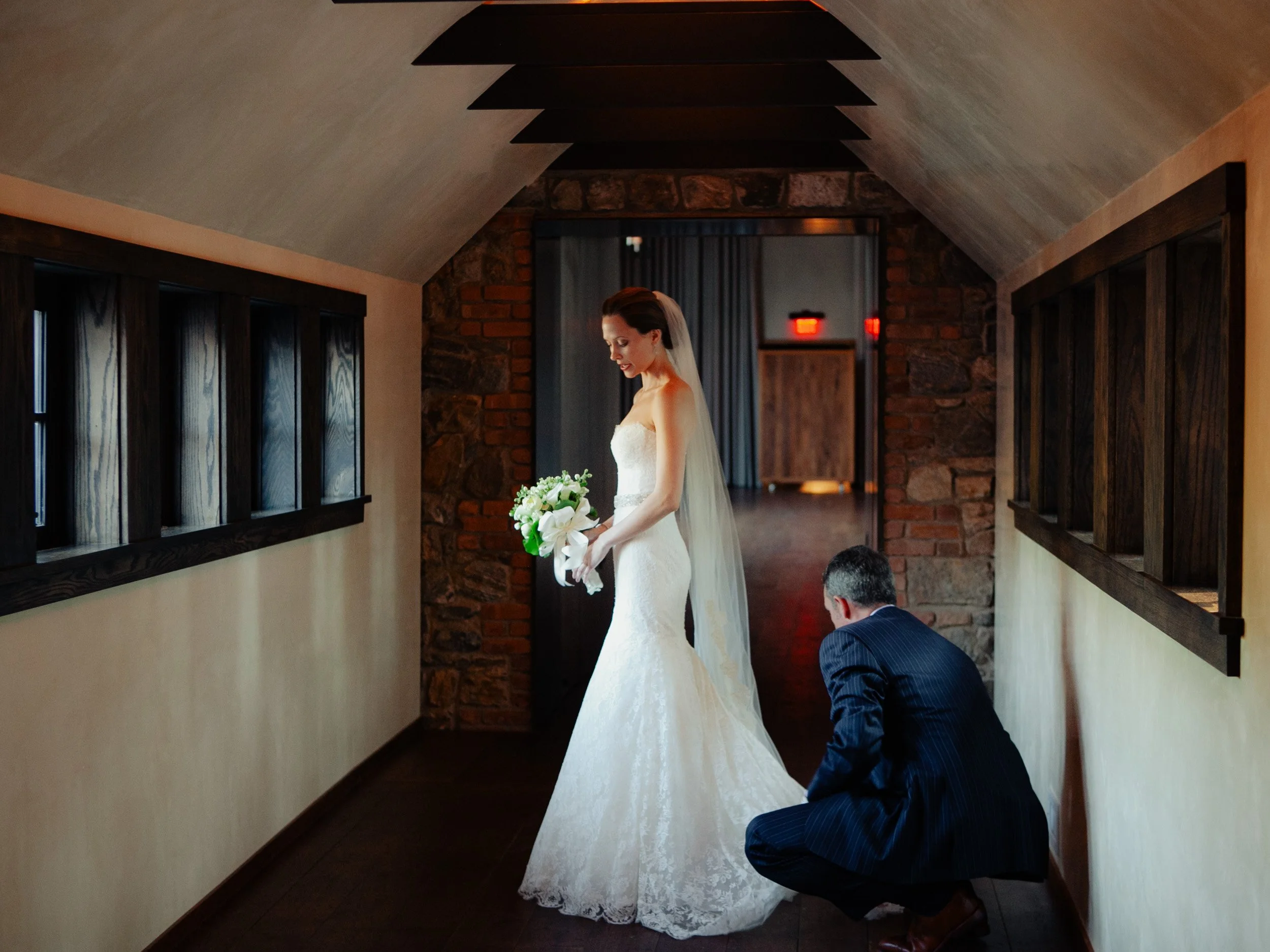 Bride in a white wedding gown holding a bouquet while a man in a suit adjusts her dress or shoe in a hallway with dark wood and brick accents.