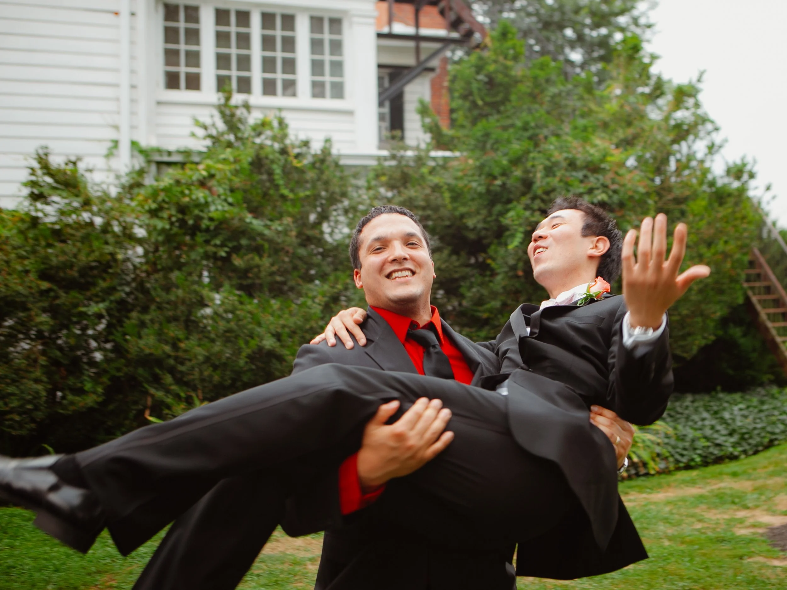 Two men in tuxedos, one is carrying the other in his arms outdoors in a garden, smiling and making a handshake gesture, with a house and greenery in the background.