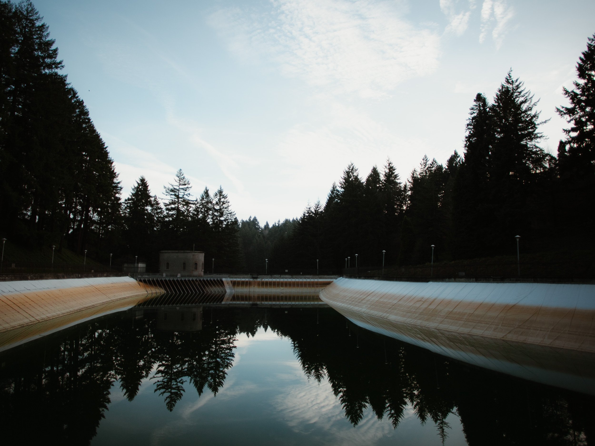 A dam with a concrete spillway, surrounded by a forest with tall trees, under a cloudy sky, reflecting in the water behind the dam.