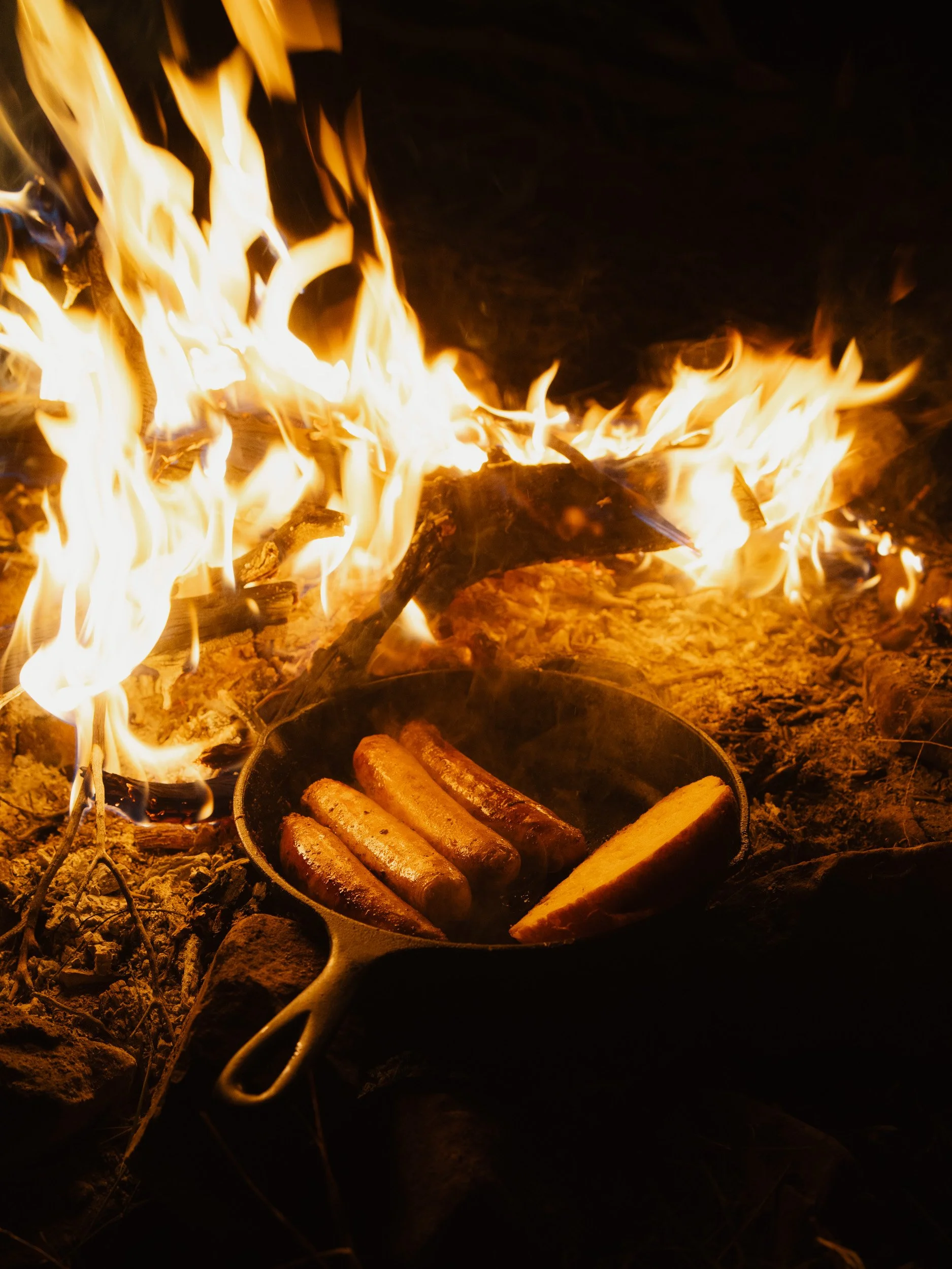 Cast iron skillet with sausages and a roasted potato on the ground in front of a campfire.