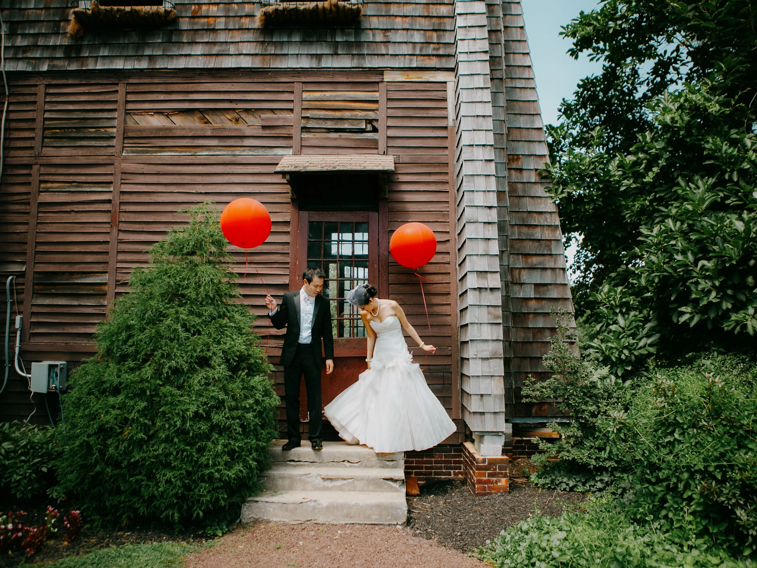 A bride and groom dressed in wedding attire standing on outdoor steps in front of a rustic wooden building, holding red balloons and leaning towards each other.
