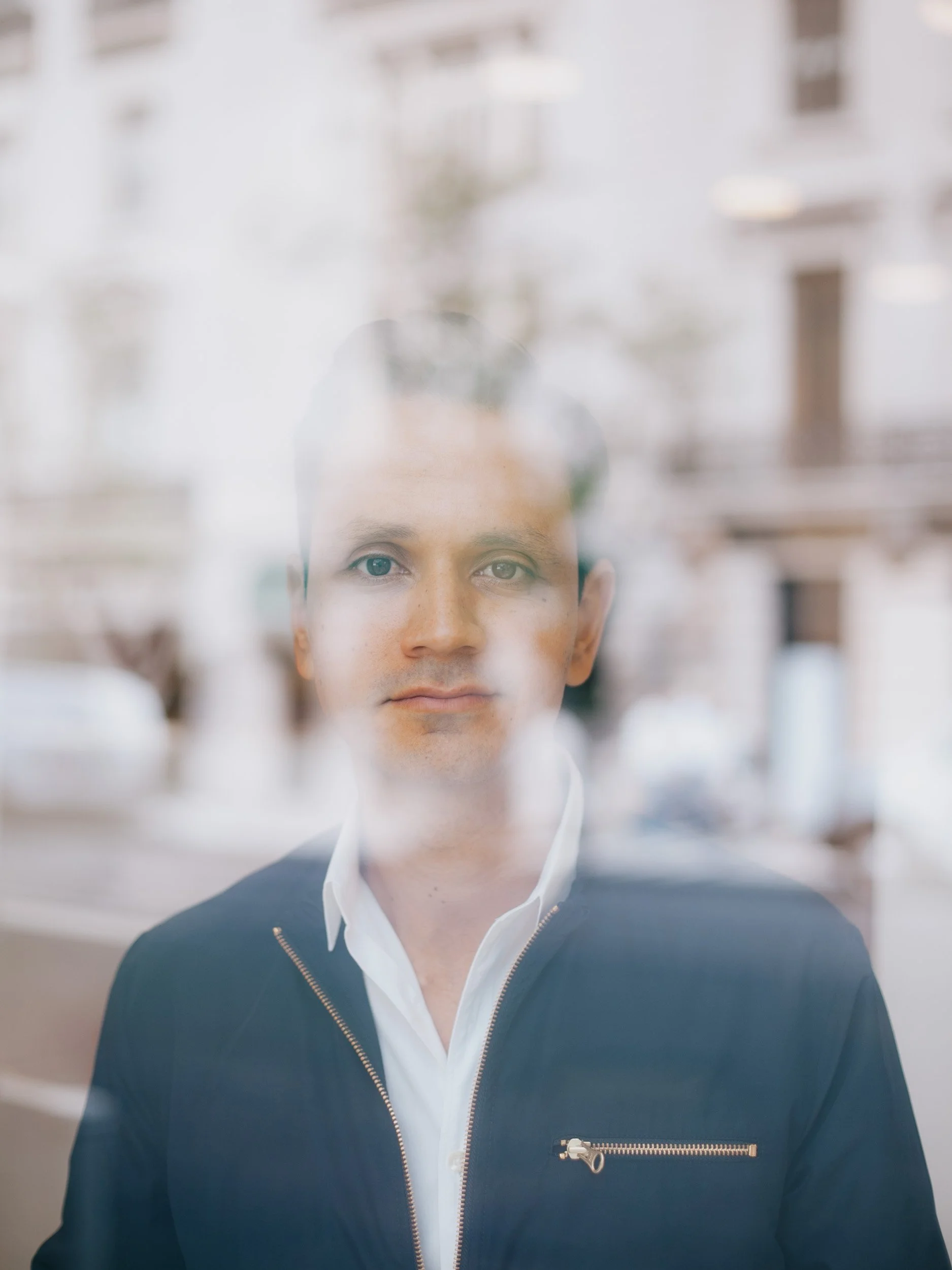 Man with short dark hair and light skin standing behind a glass window or door with reflections, wearing a white collared shirt and black zip-up jacket.