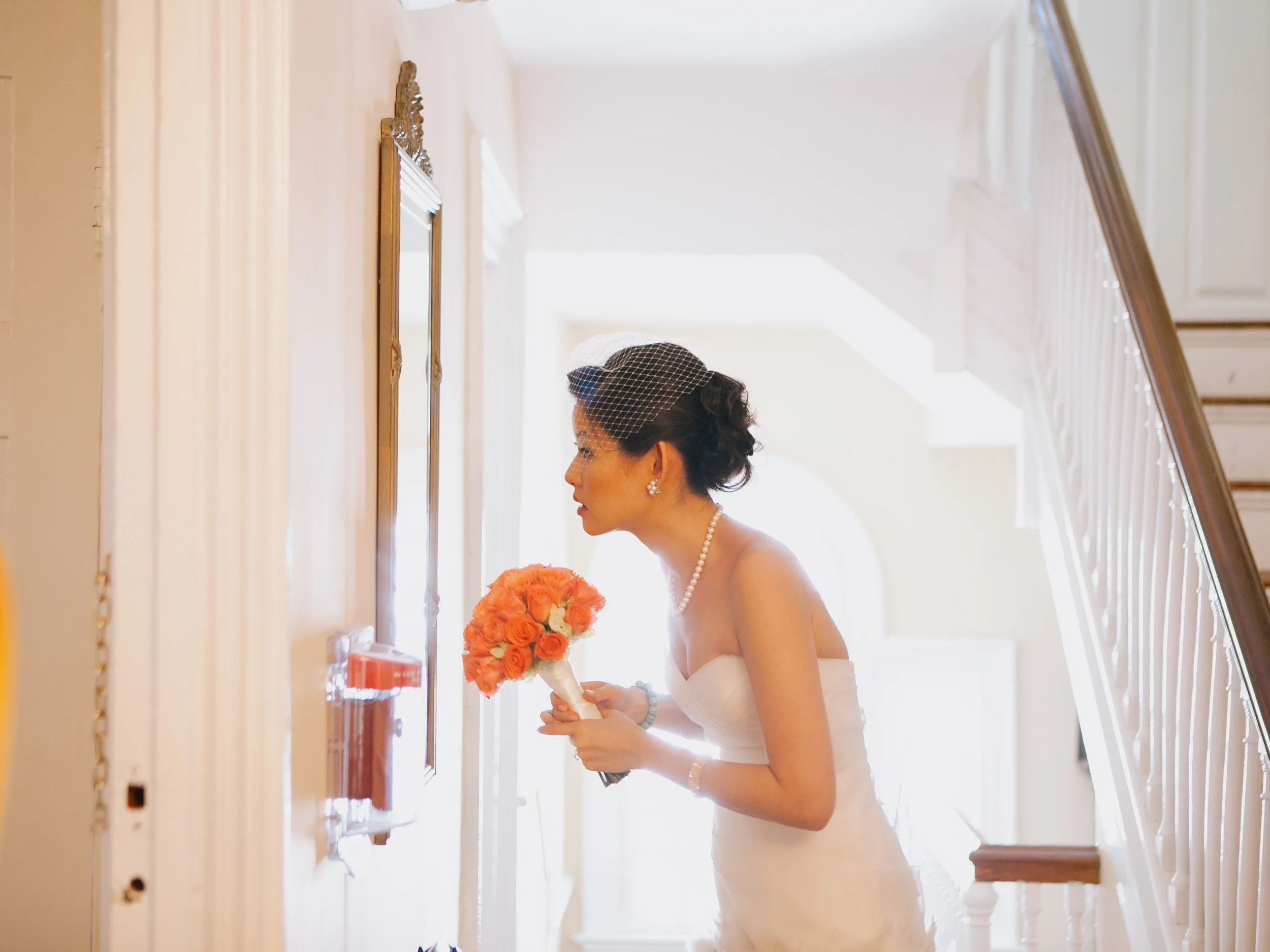 A bride in a strapless white wedding dress and pearl necklace looks through a window on a staircase landing, holding a bouquet of orange roses, with a black veil over her hair.