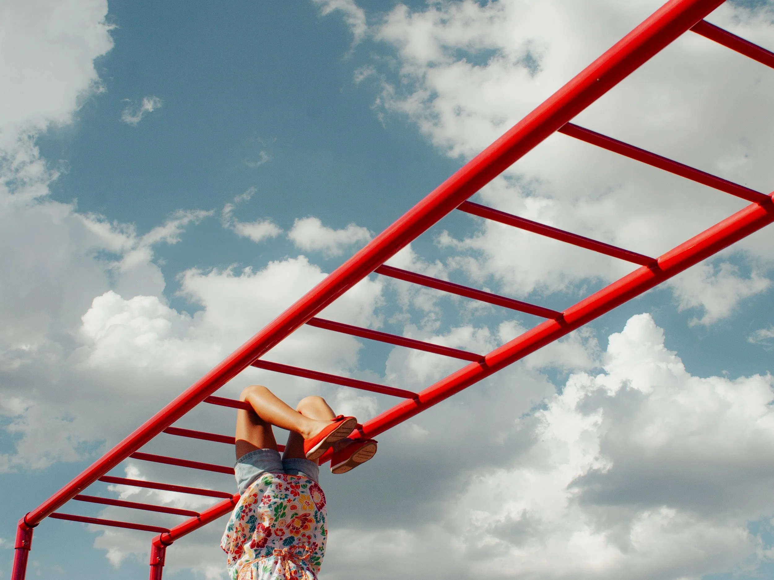 Child hanging upside down from a red jungle gym outdoors against a blue sky with clouds.