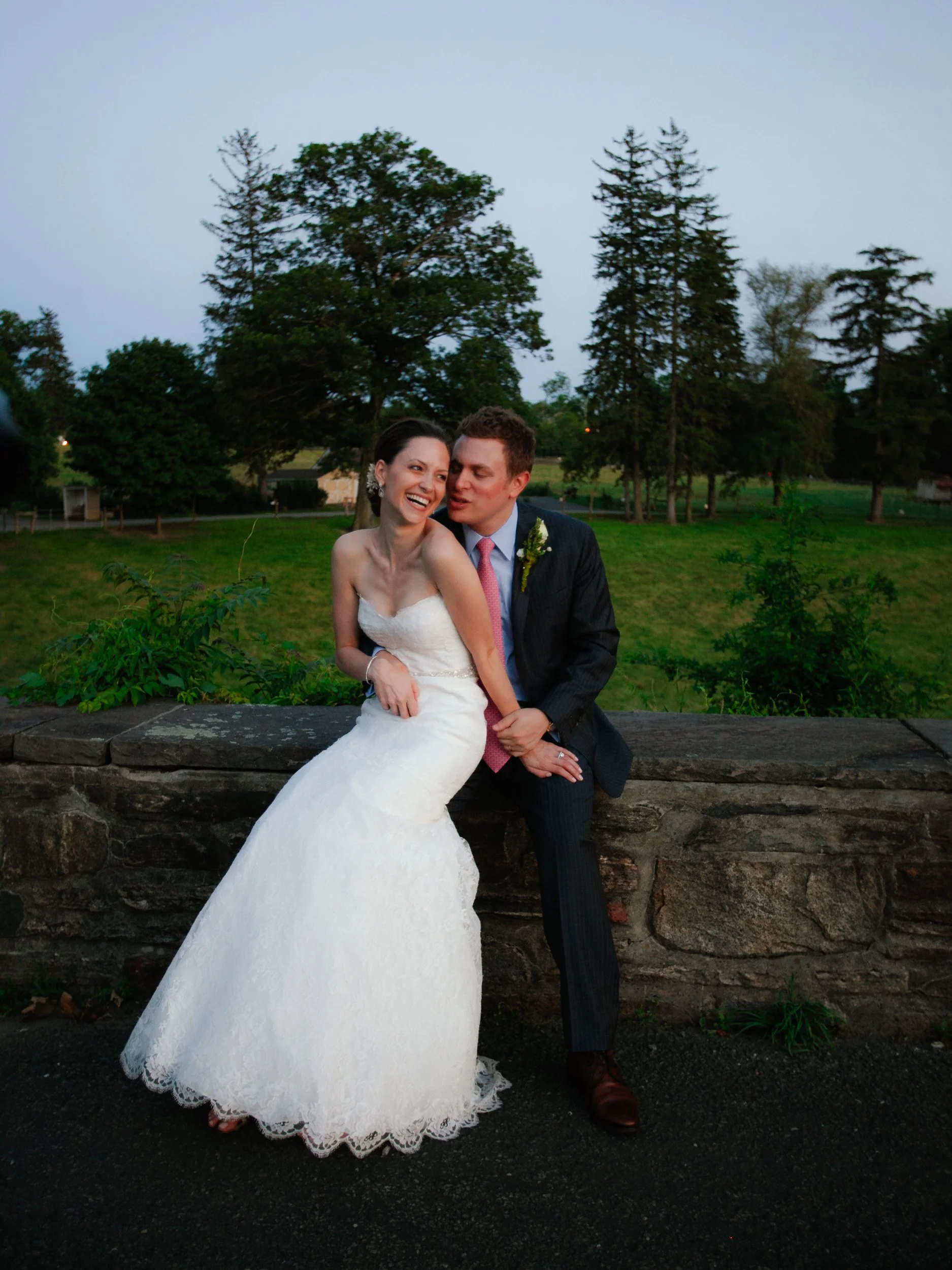 A bride and groom sitting on a stone wall outdoors, smiling and laughing, with trees and a grassy field in the background during a wedding photo.