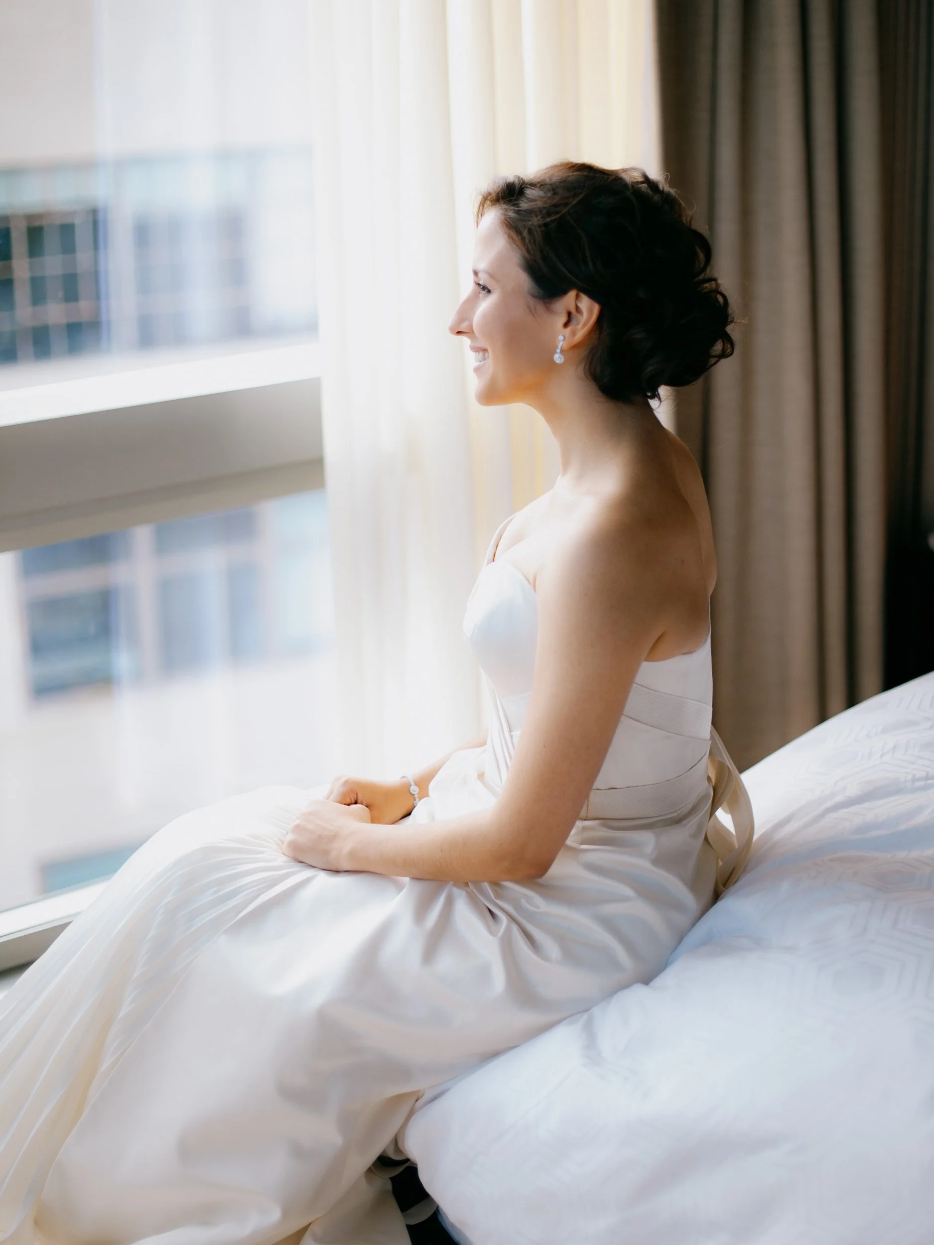 A woman in a white dress sitting on a bed, looking out a window with a soft smile.
