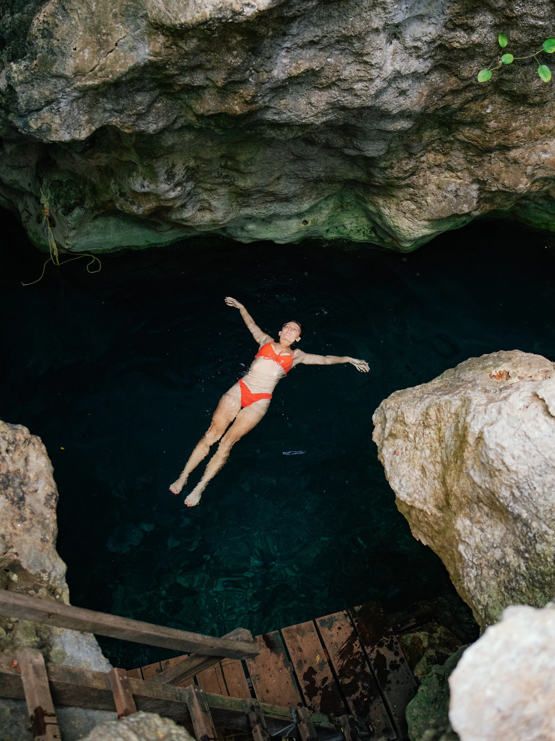 A woman in a red bikini floating in a deep, dark natural water cave surrounded by rocks.