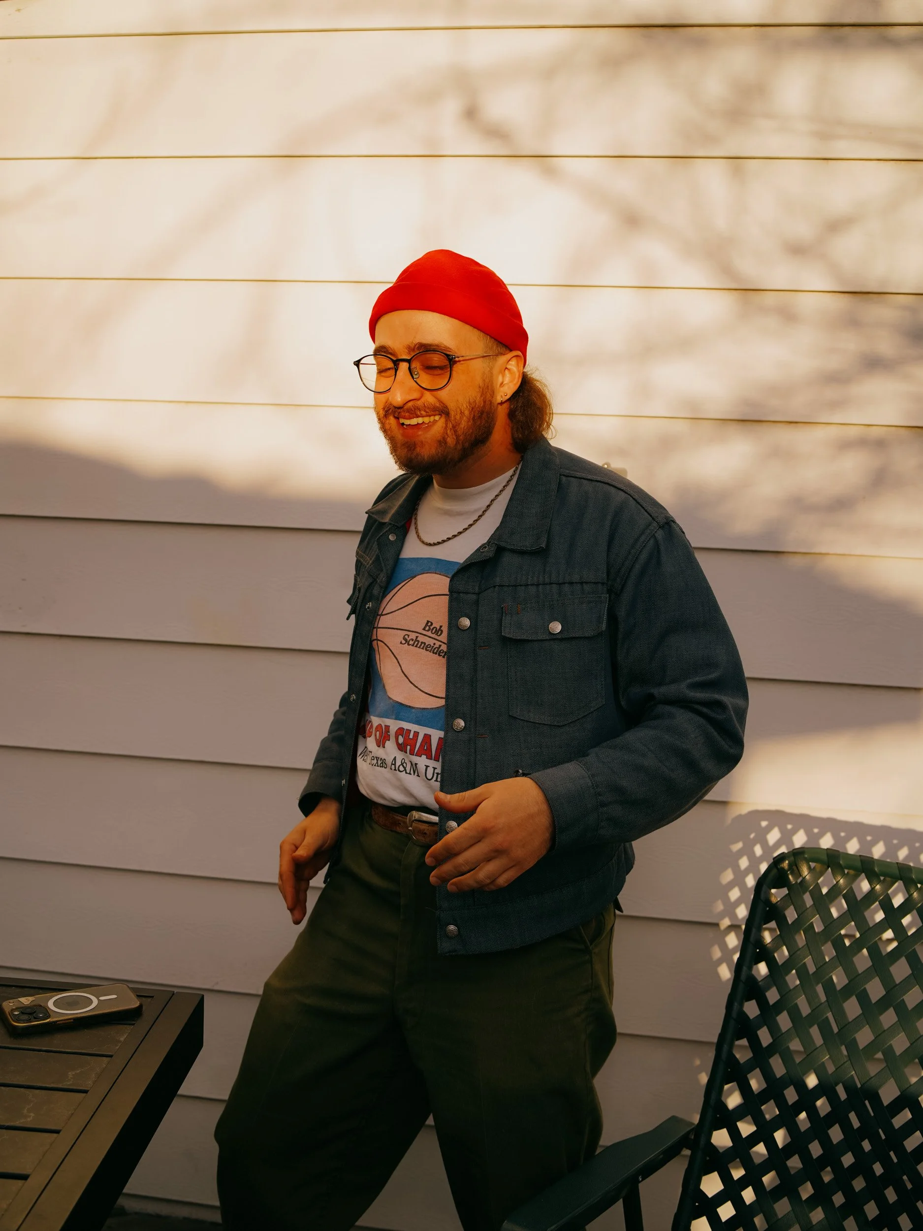 A man with glasses, a beard, and long hair wearing a red beanie, denim jacket, and a graphic T-shirt, smiling outdoors next to a white wall and a black chair.
