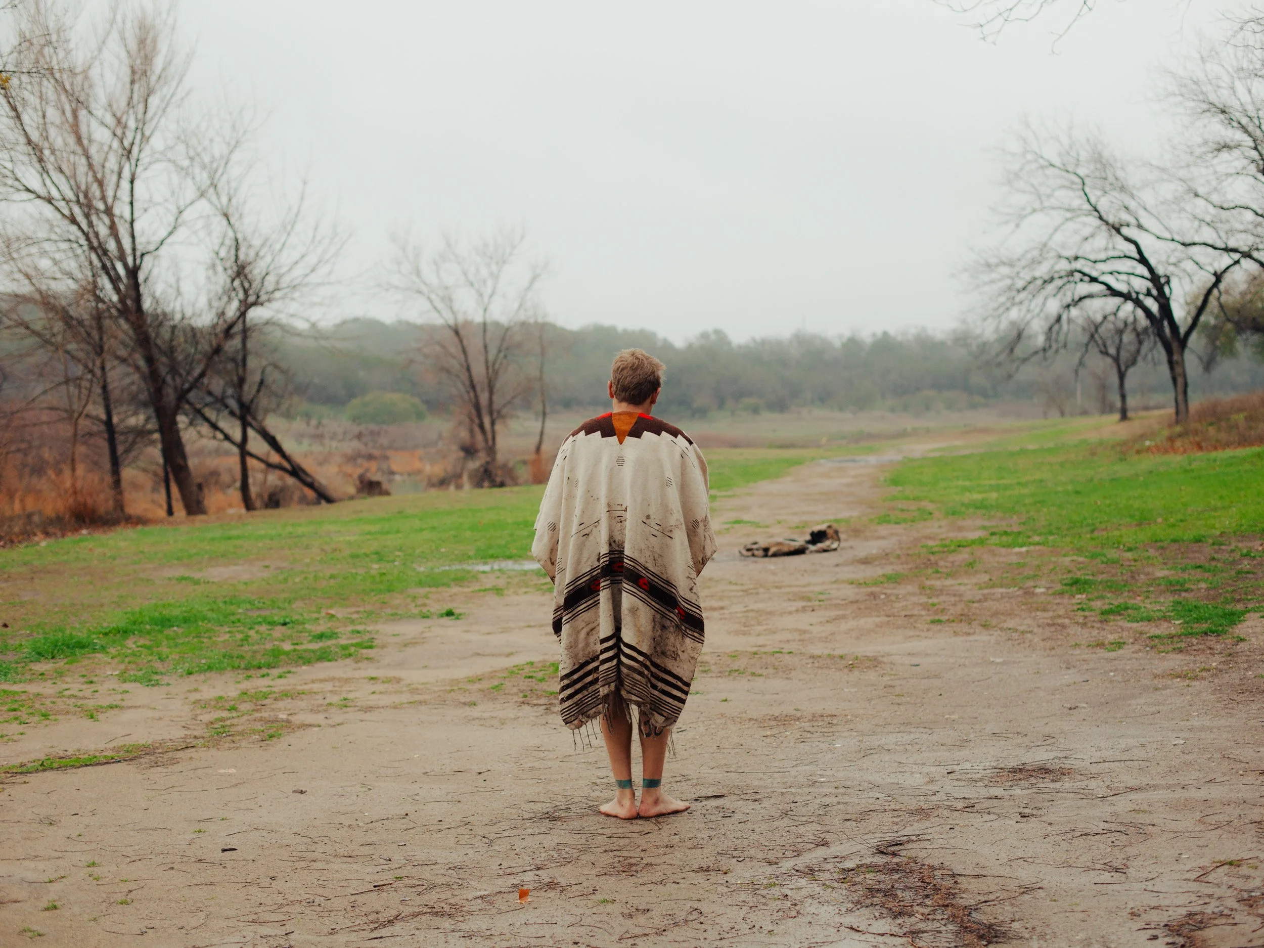 Person wrapped in a blanket walking barefoot on a dirt path in a park with leafless trees and green grass, overcast sky.